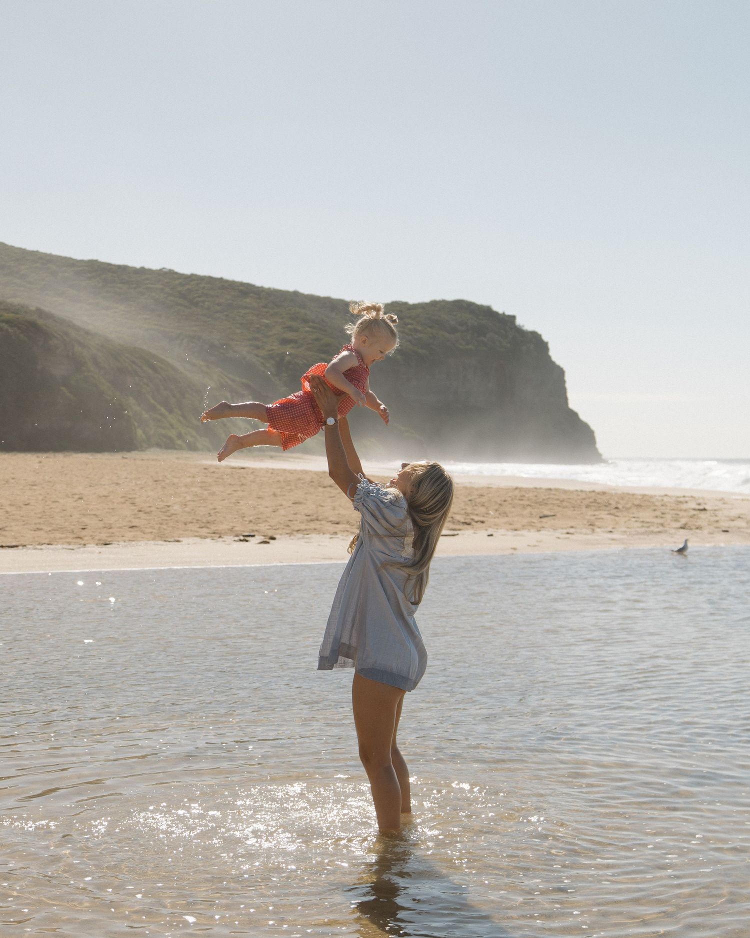 A mother joyfully lifts her baby in the air while standing in shallow beach water with a scenic cliff backdrop.