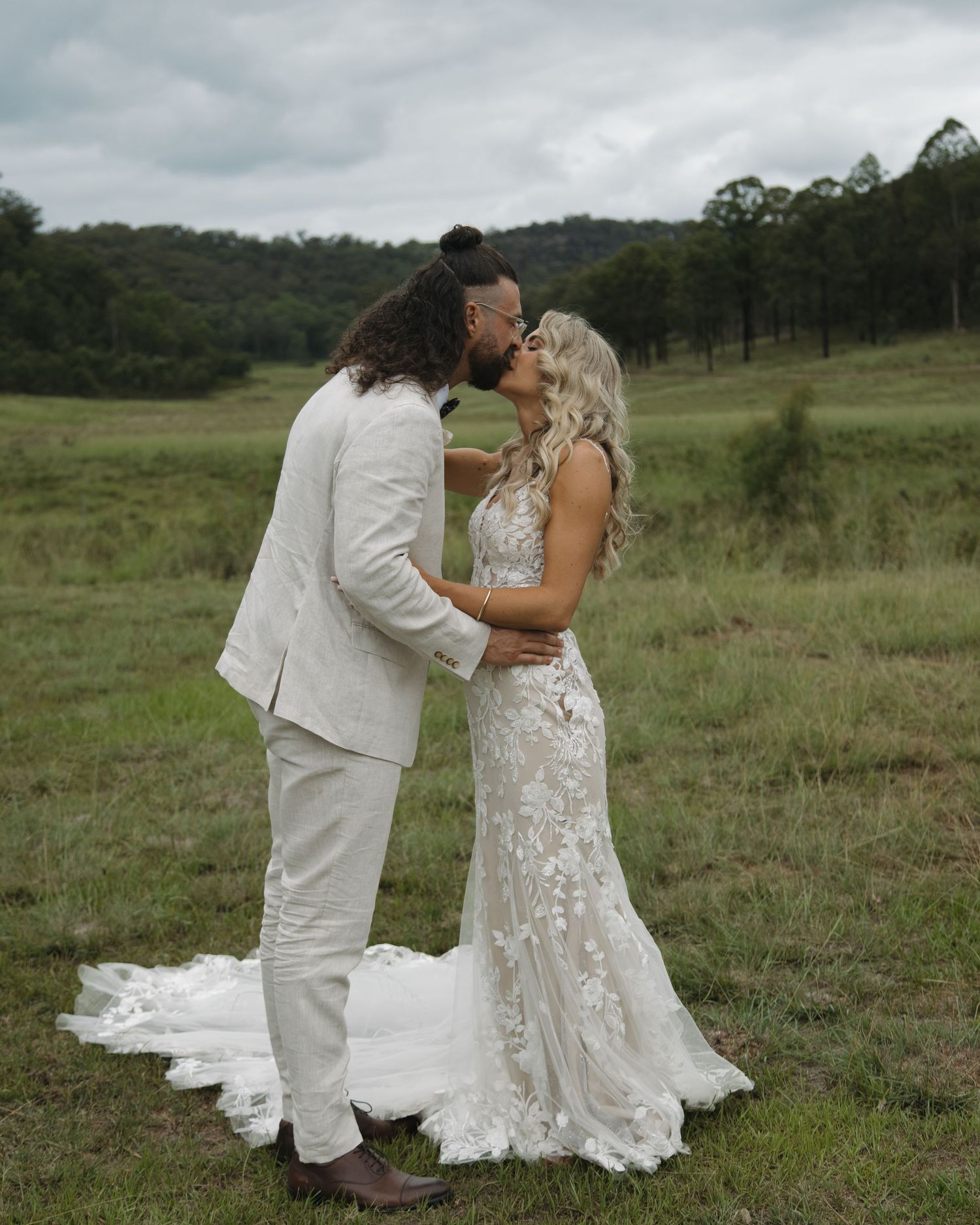 A bride and groom share a romantic kiss in a lush green field, wearing elegant white wedding attire.