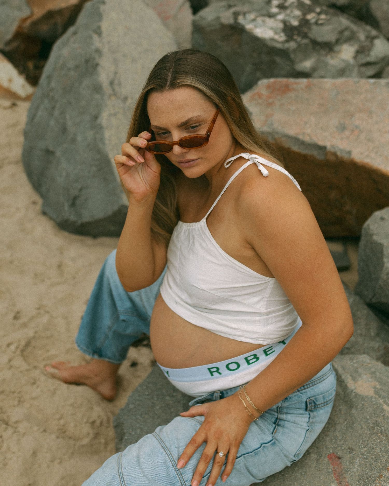 Person in white crop top and jeans poses on rocky beach setting.
