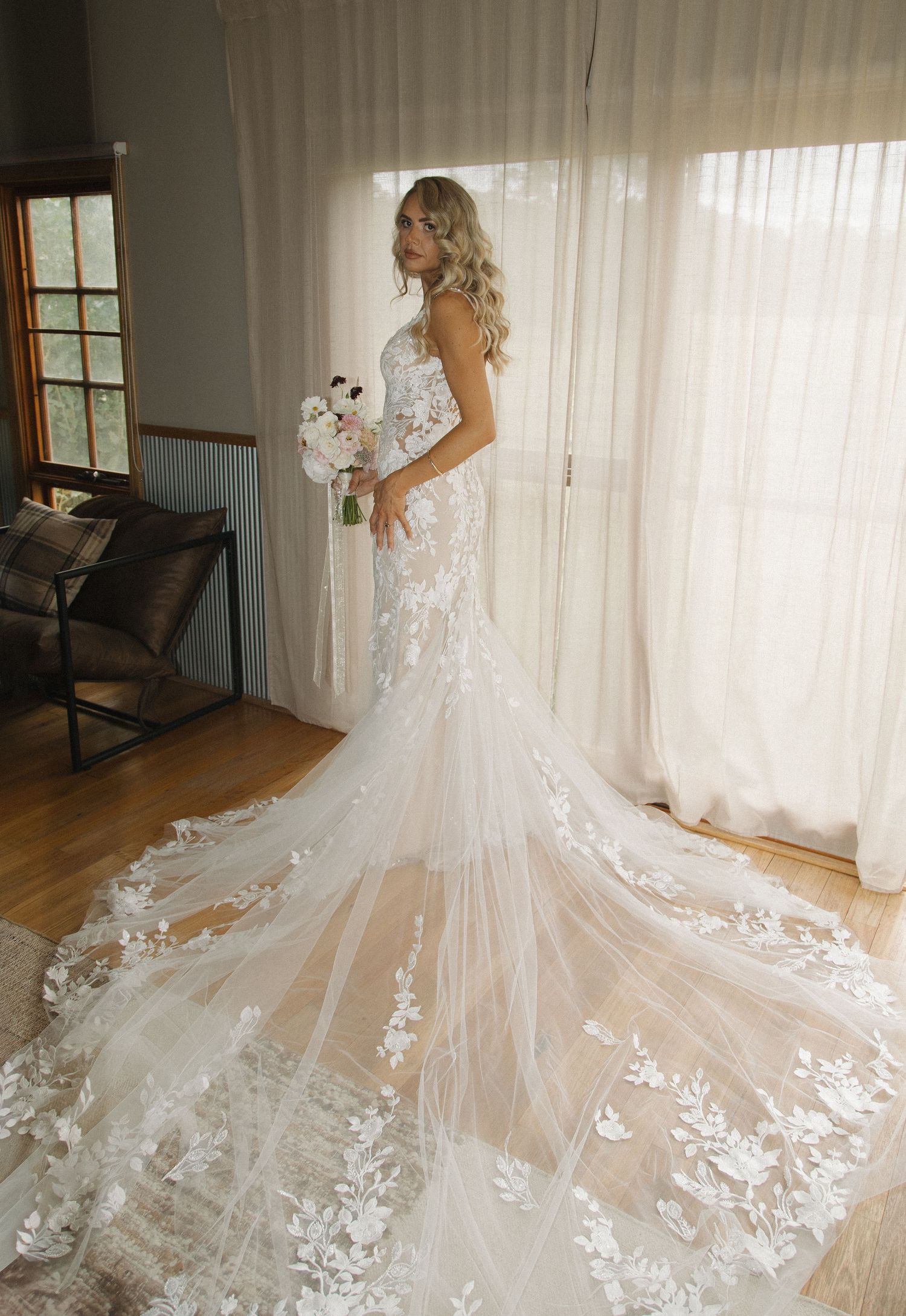 Bride in elegant lace mermaid wedding gown with floral appliques and cathedral train, holding bouquet by window.