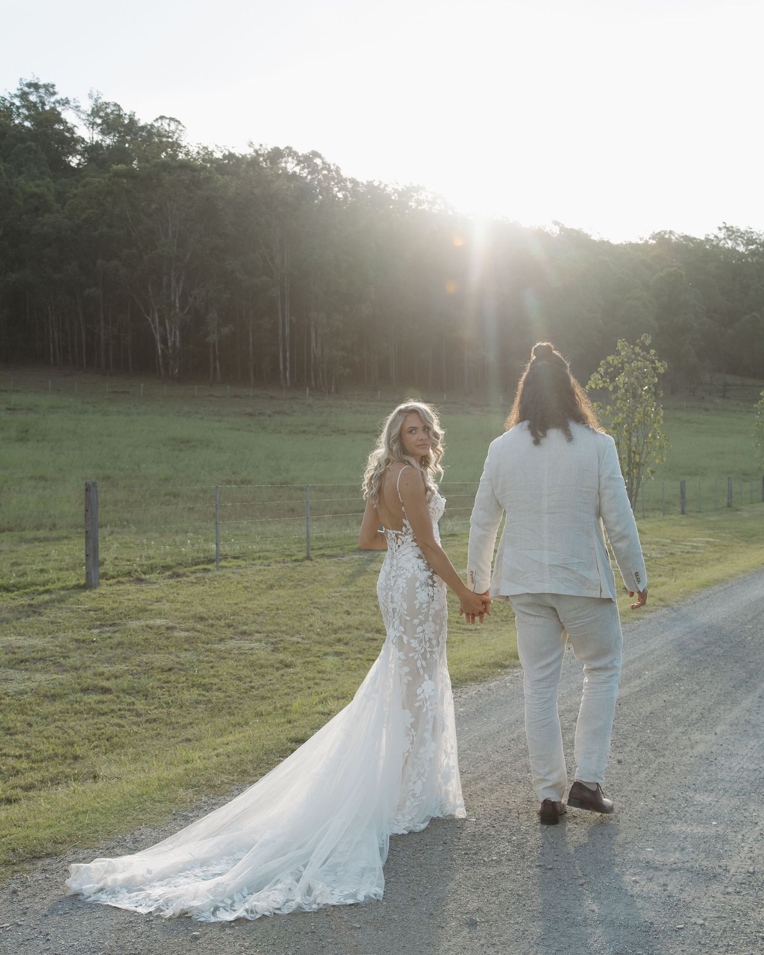 Bride and groom holding hands walking down a sunlit country road at golden hour, with lush green fields and trees in background.