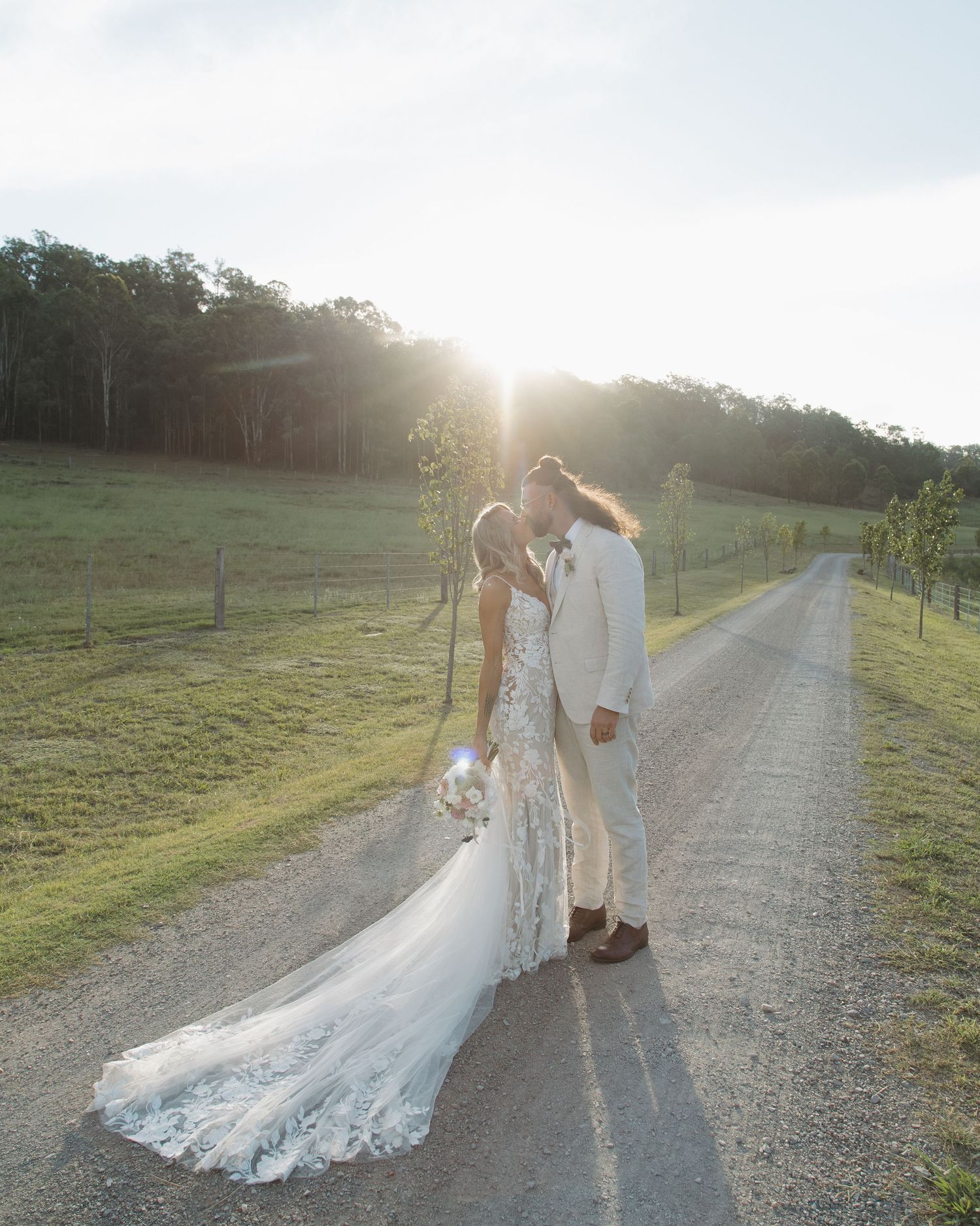 A bride and groom share a romantic kiss at golden hour on a country road, surrounded by lush green fields.
