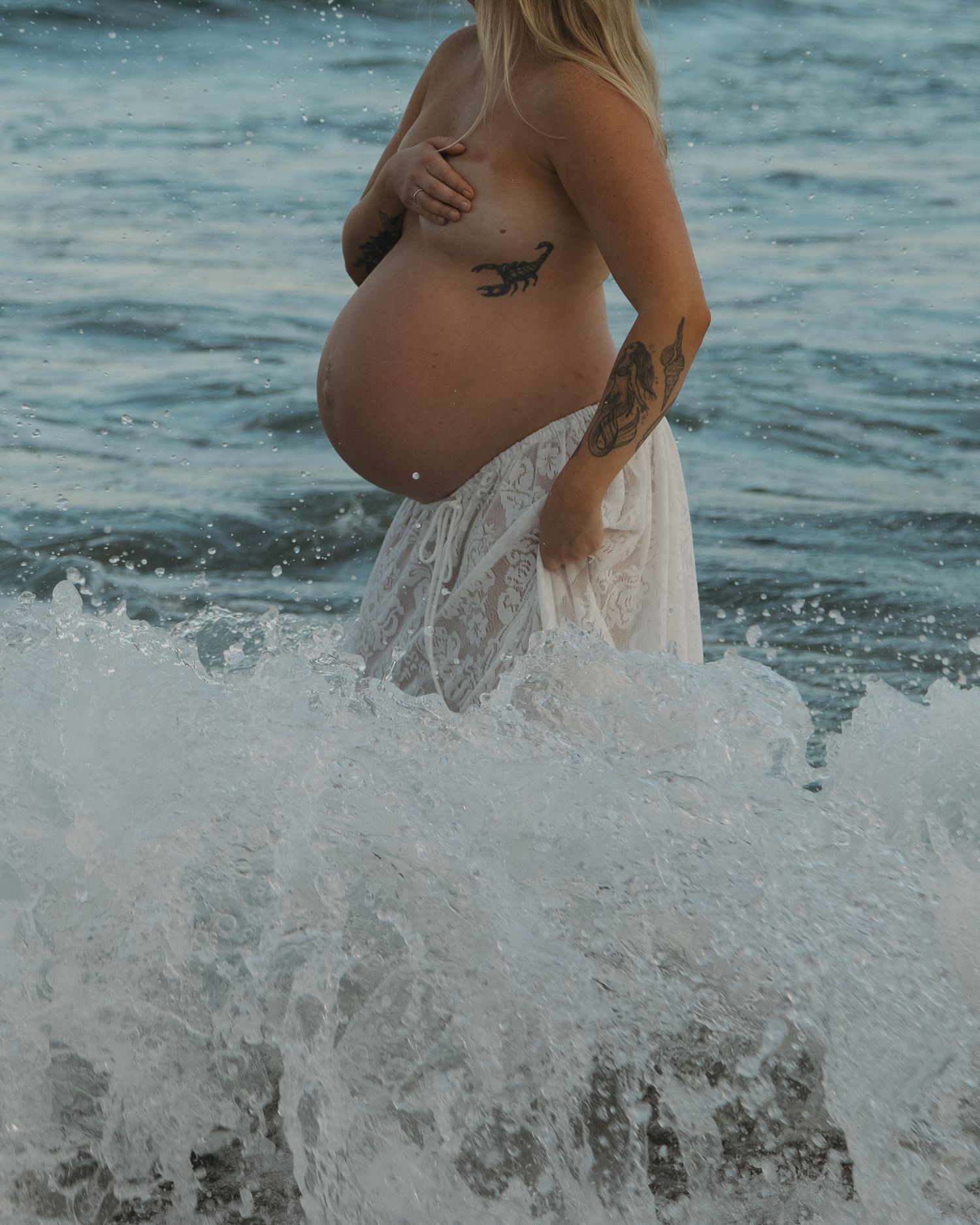 A pregnant figure stands in flowing white fabric as ocean waves crash around them at the beach.