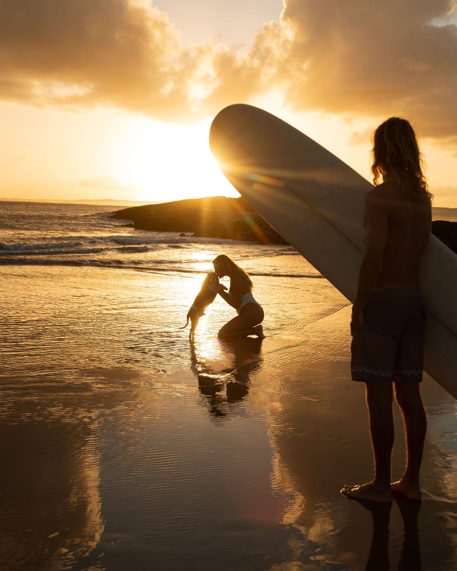 A silhouetted figure with a surfboard stands on a beach at sunset while golden light illuminates the ocean waves.