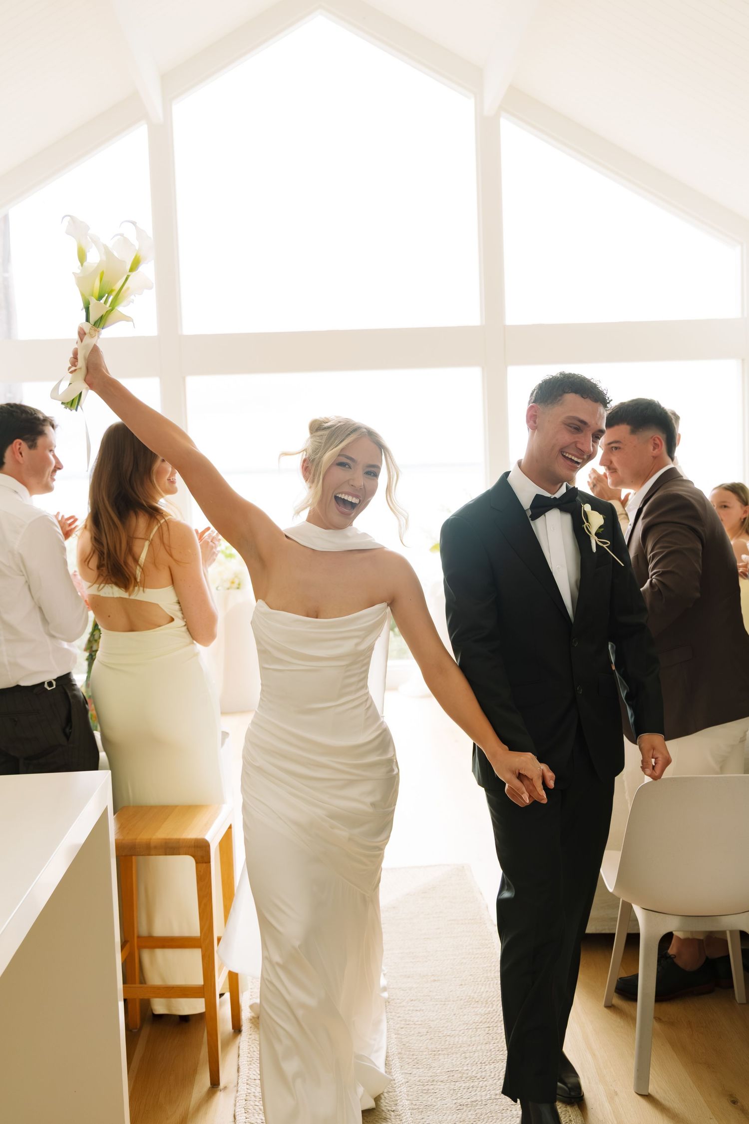 Joyful bride in white gown raises bouquet while groom in tuxedo holds her hand after wedding ceremony.