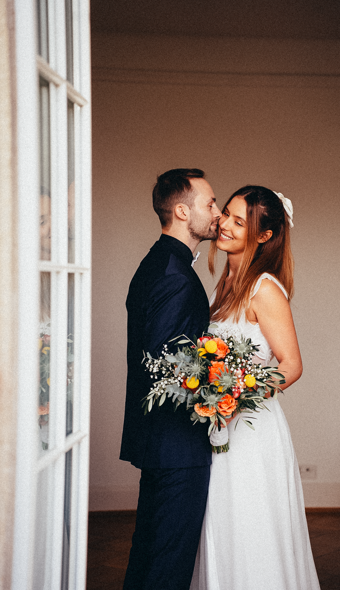Groom kisses smiling bride on cheek as she holds vibrant orange and yellow wedding bouquet indoors.