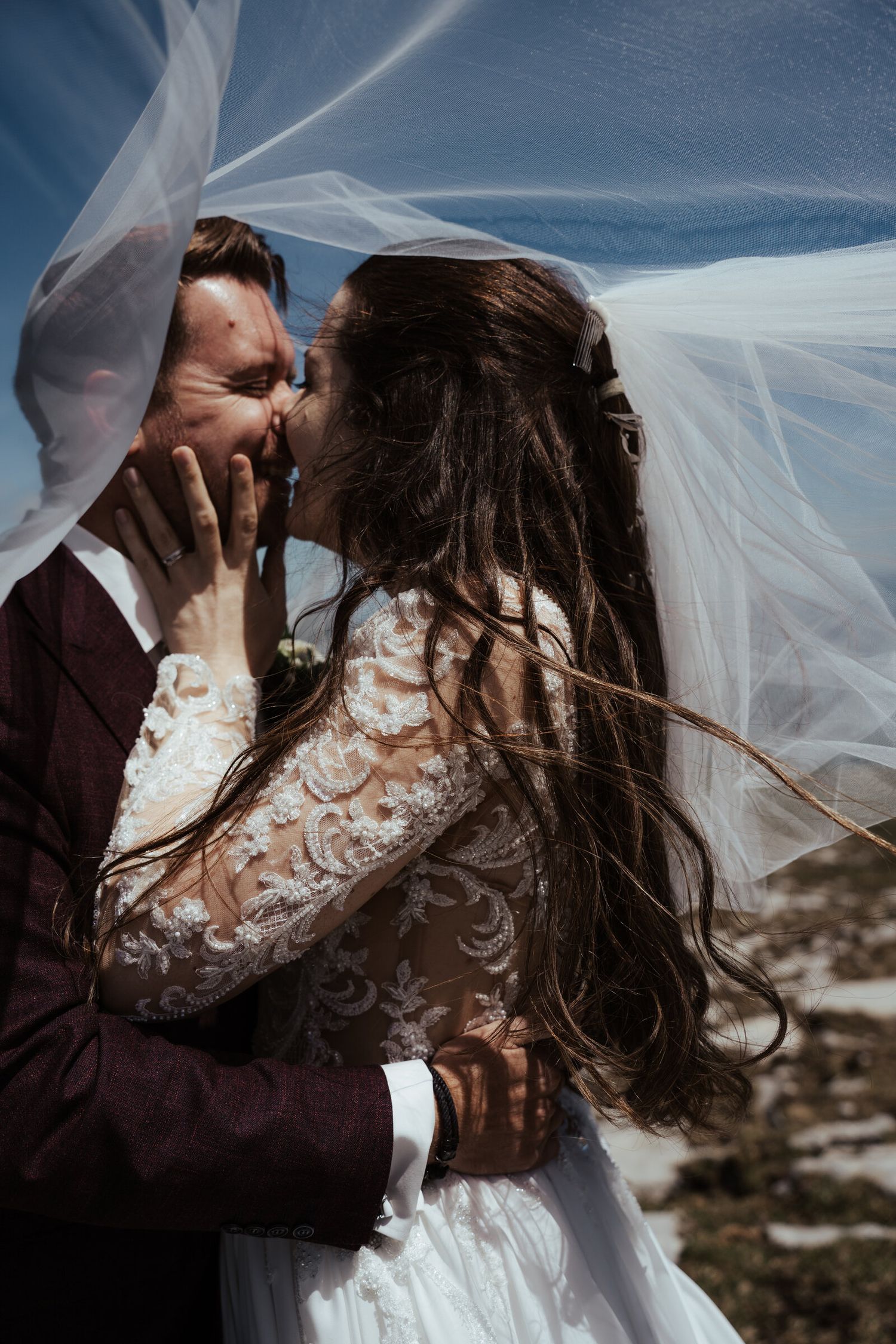 Bride and groom sharing a romantic kiss under a flowing veil outdoors on their wedding day.