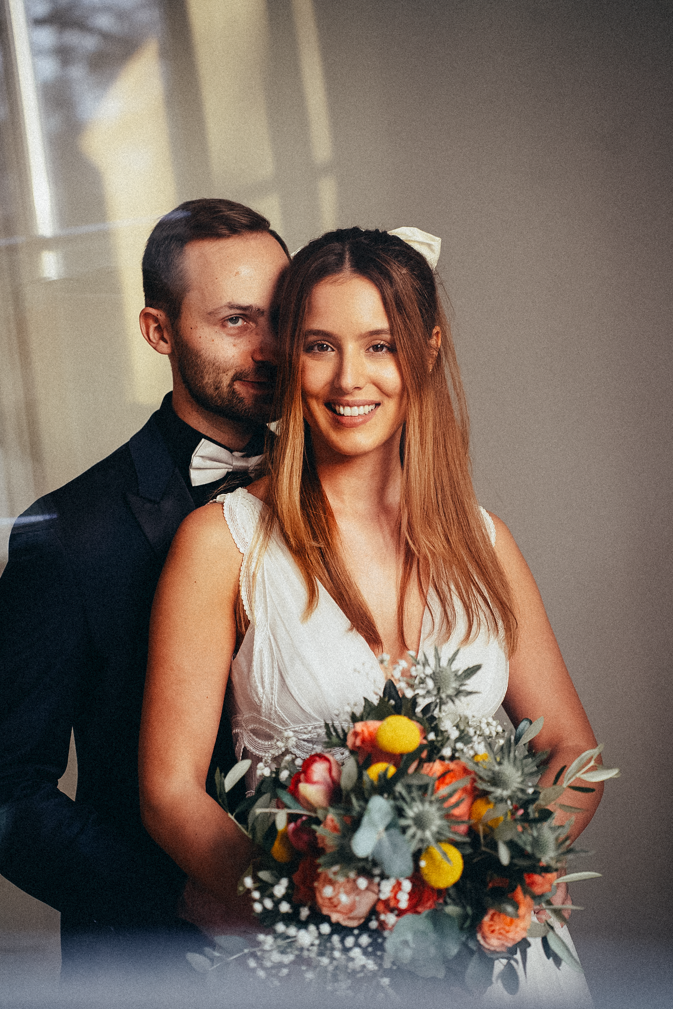 Happy bride smiles holding vibrant bouquet while groom embraces her from behind near a sunlit window.