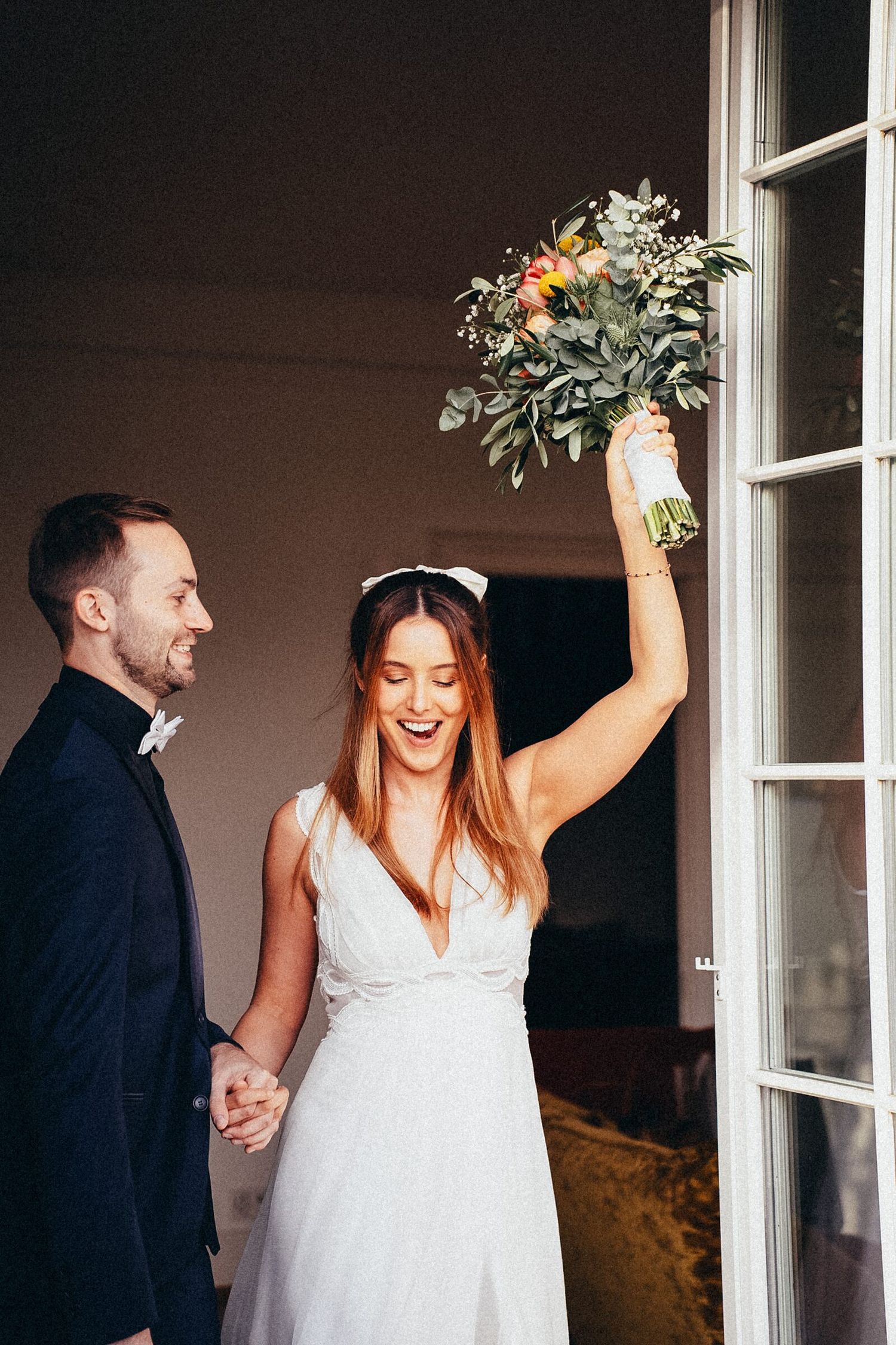 Joyful bride in white dress raises bouquet while groom in navy suit holds her hand at wedding venue.