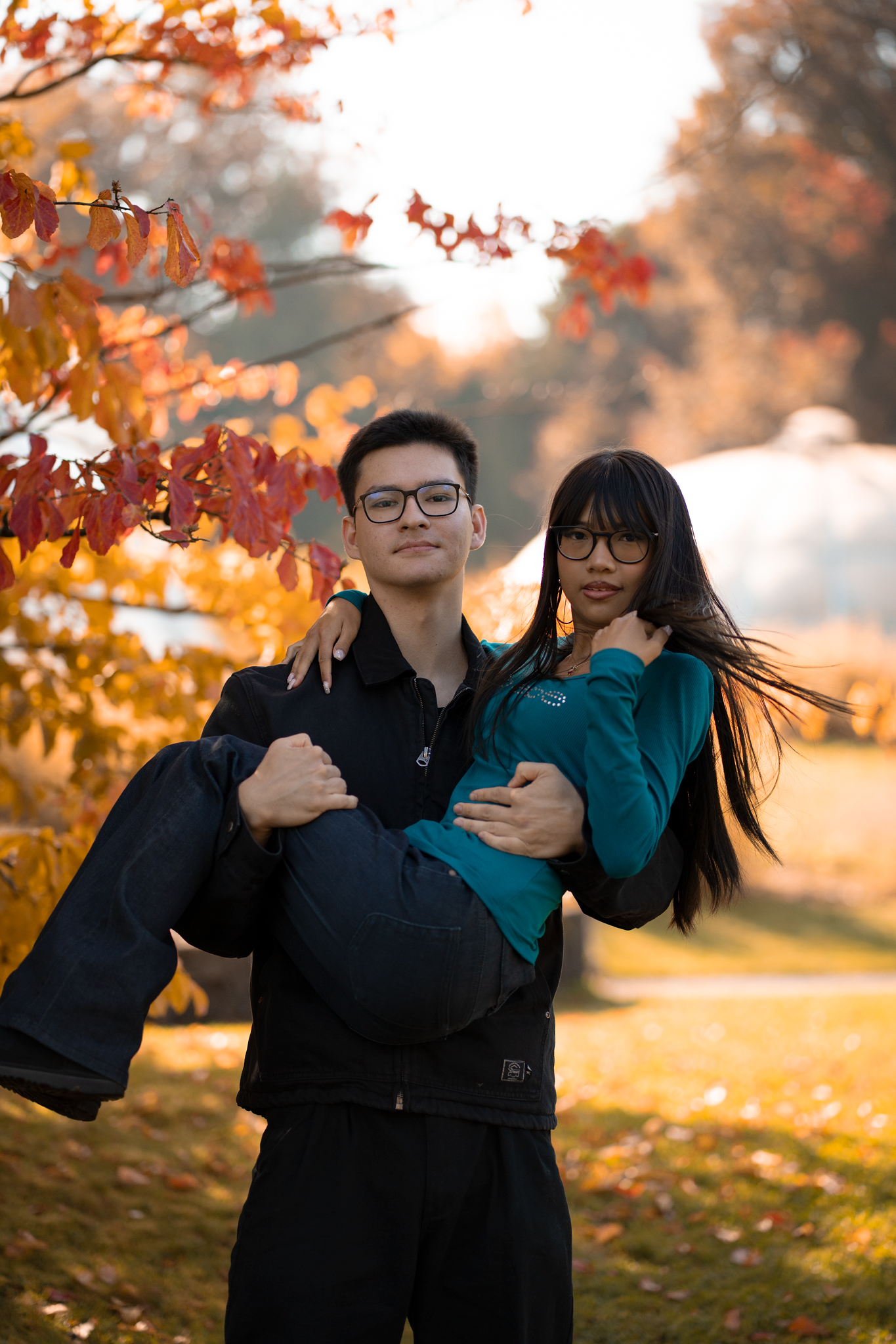 A young man carries a woman in his arms among vibrant autumn foliage in a sunlit park.