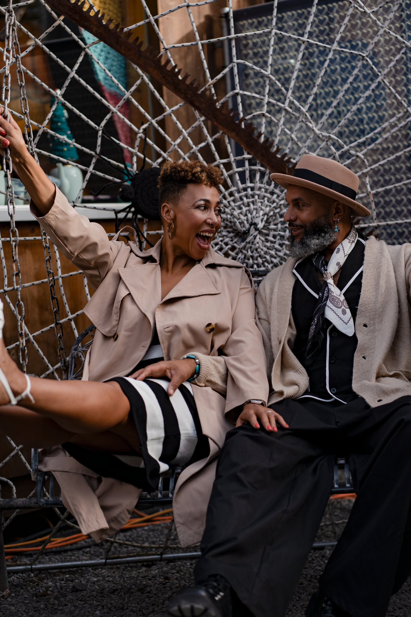 Two stylish people laughing and sitting together in front of a decorative metal backdrop at an outdoor event.