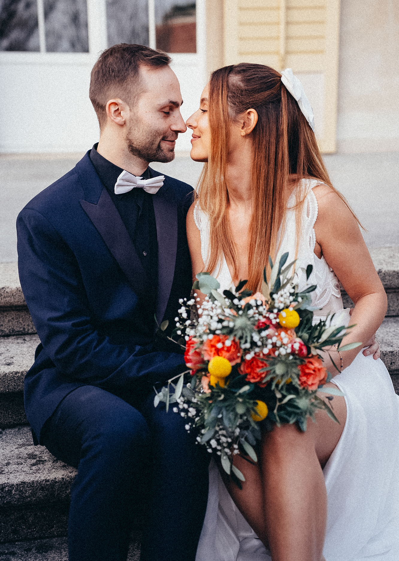 Bride and groom sitting on steps, sharing an intimate moment, with a colorful bouquet of red and yellow flowers.