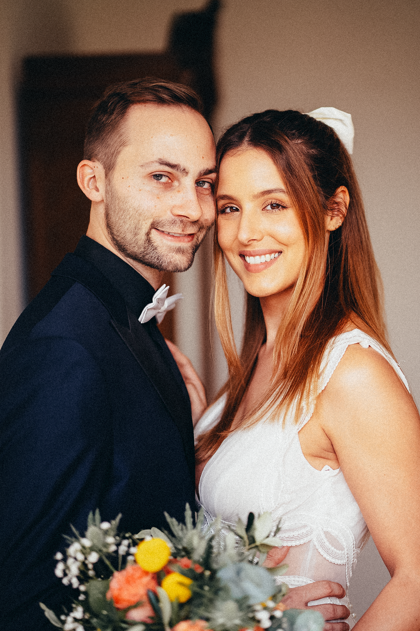 Smiling bride and groom in wedding attire pose together, she holds a colorful bouquet with orange and yellow flowers.