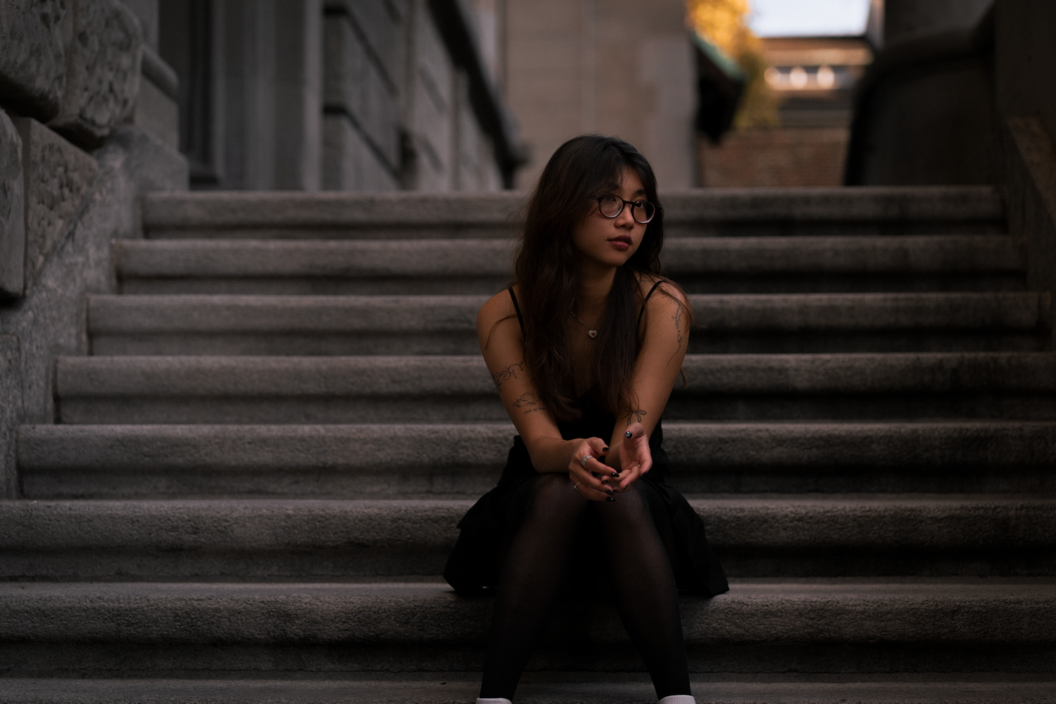 Young woman with glasses and tattoos sits thoughtfully on stone steps in dim light, conveying a pensive and introspective mood.