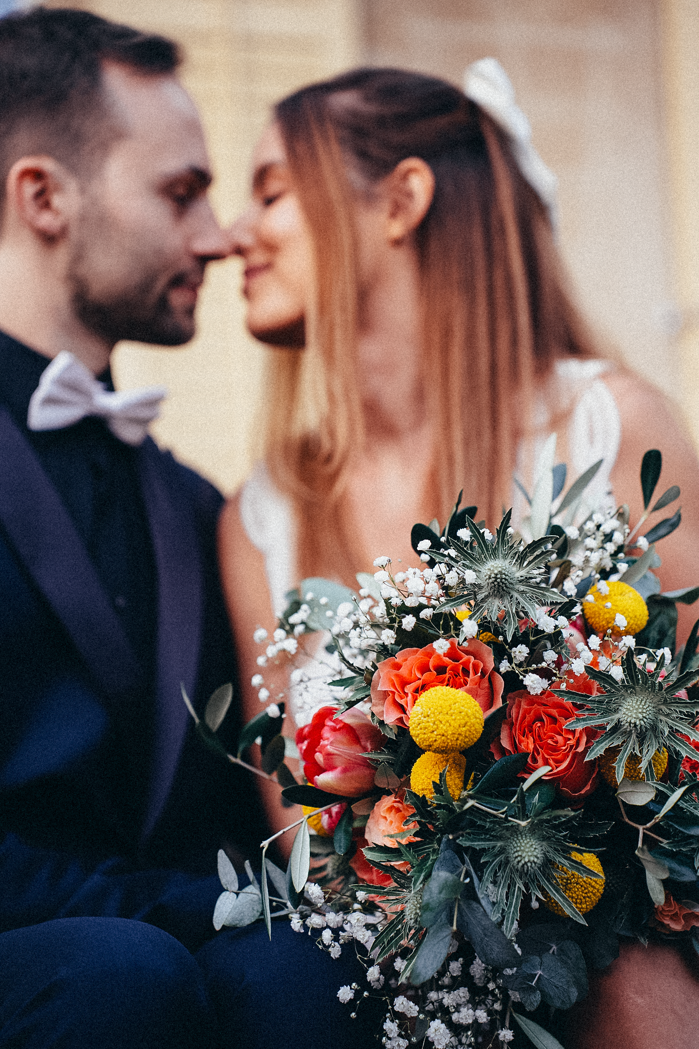 Wedding couple almost kissing with colorful bridal bouquet of orange, yellow, and white blooms in foreground.