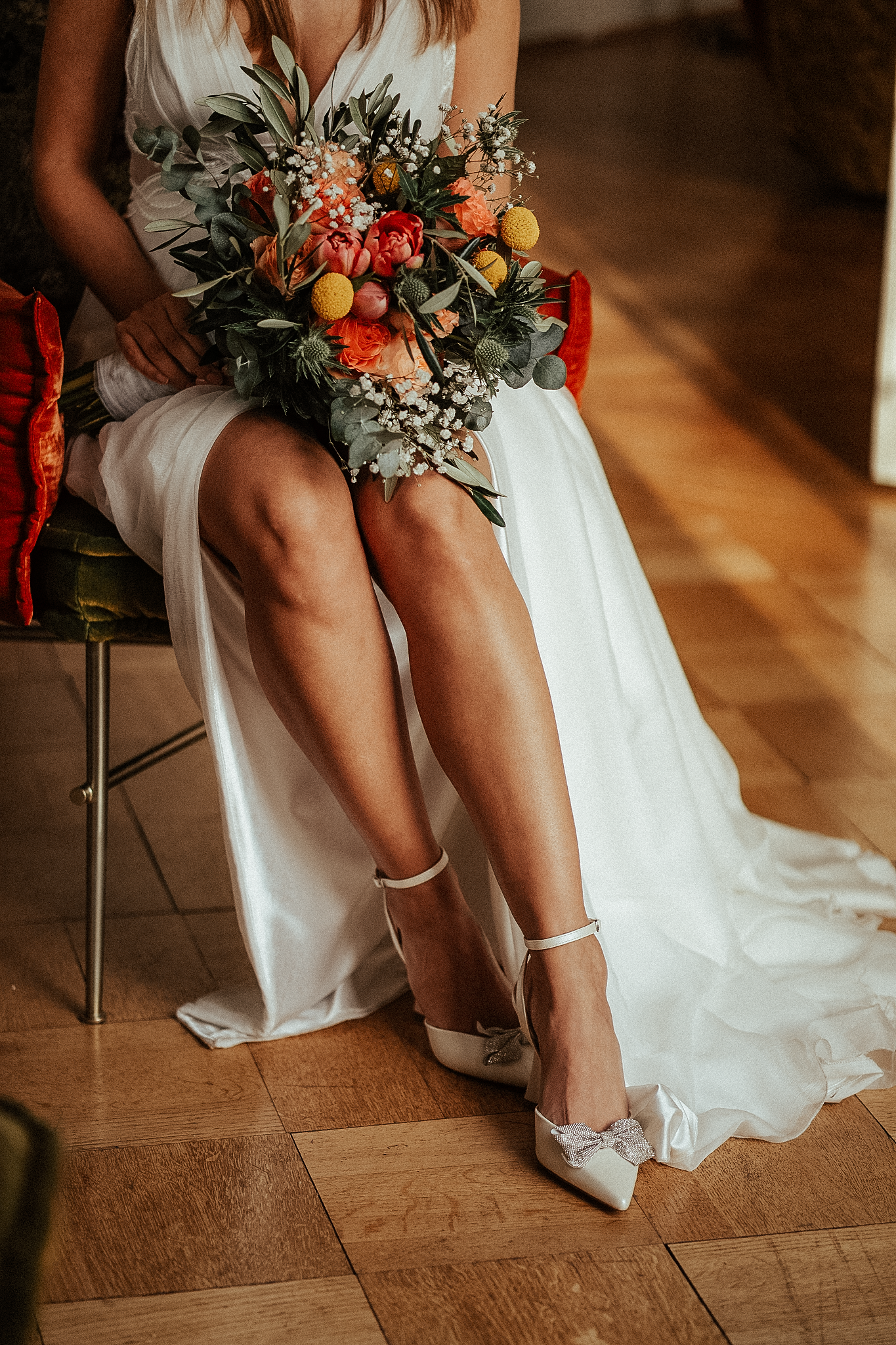 Bride in white wedding dress holding colorful floral bouquet while seated, wearing elegant heeled shoes on wooden floor.