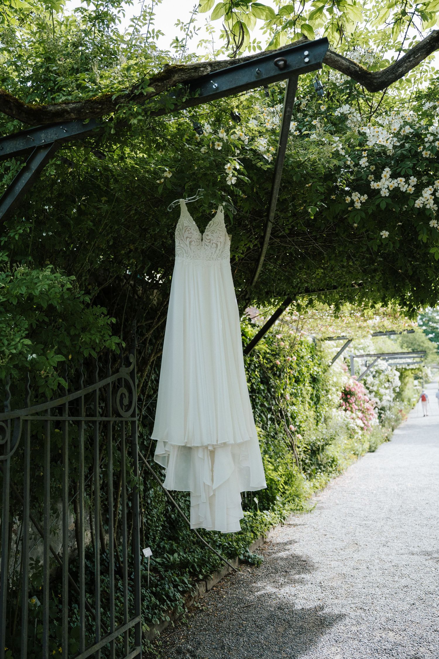 White wedding dress hanging on a metal arch in a lush green garden pathway with blooming flowers.
