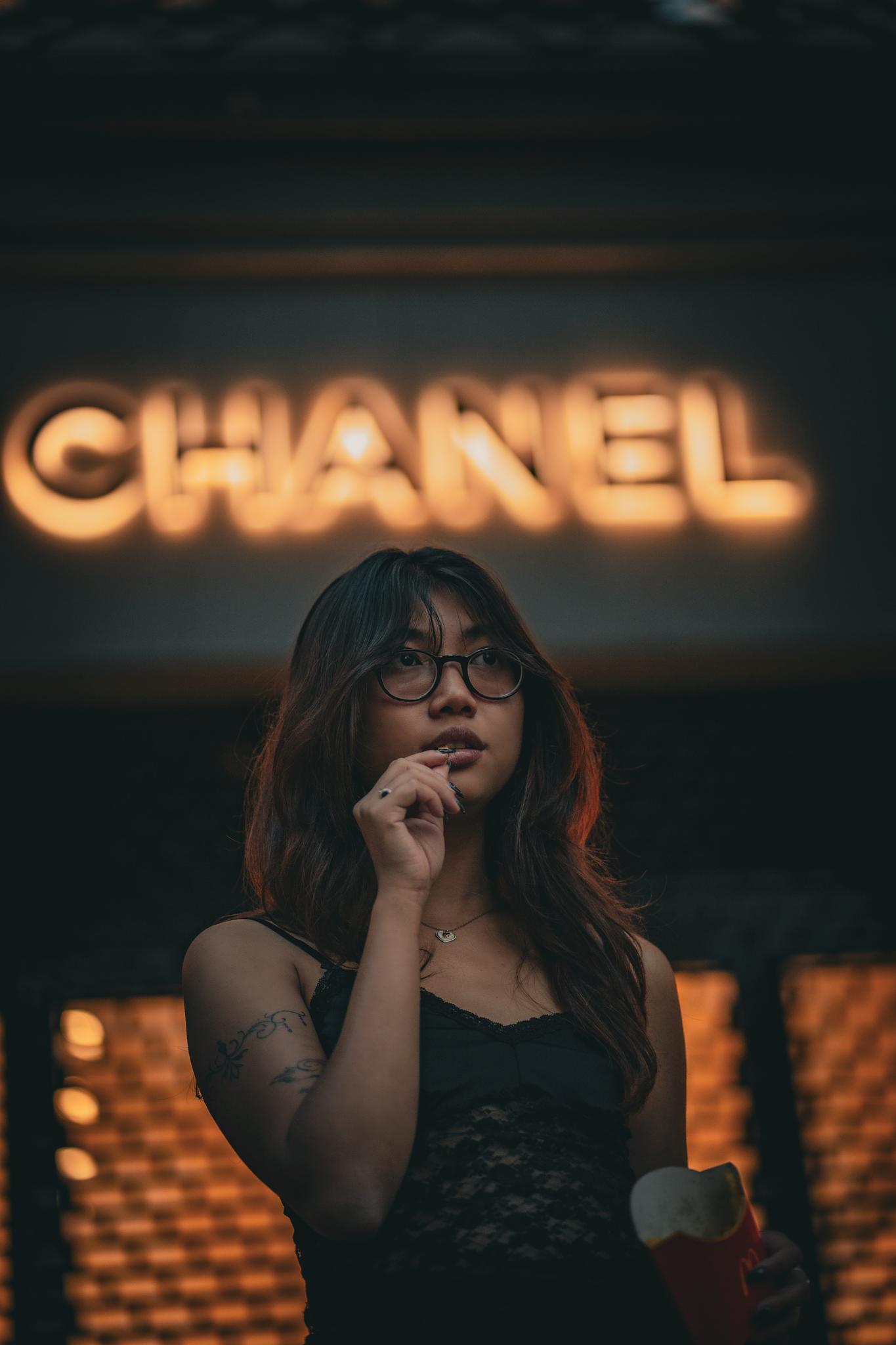 Young woman with glasses poses thoughtfully before a glowing Chanel neon sign at night.