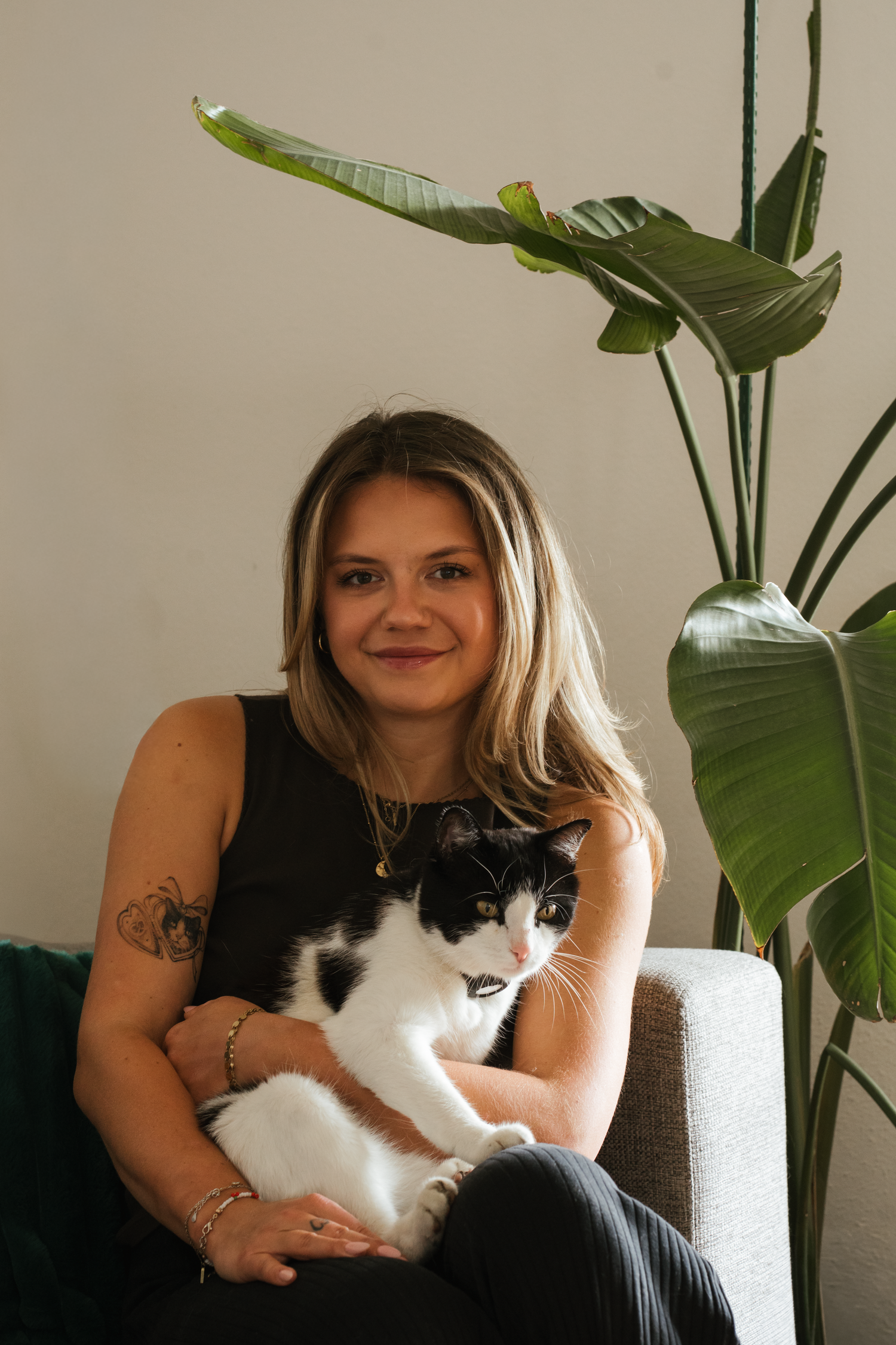Smiling blonde woman with tattoos sitting in a chair holding a black and white cat near a large plant.