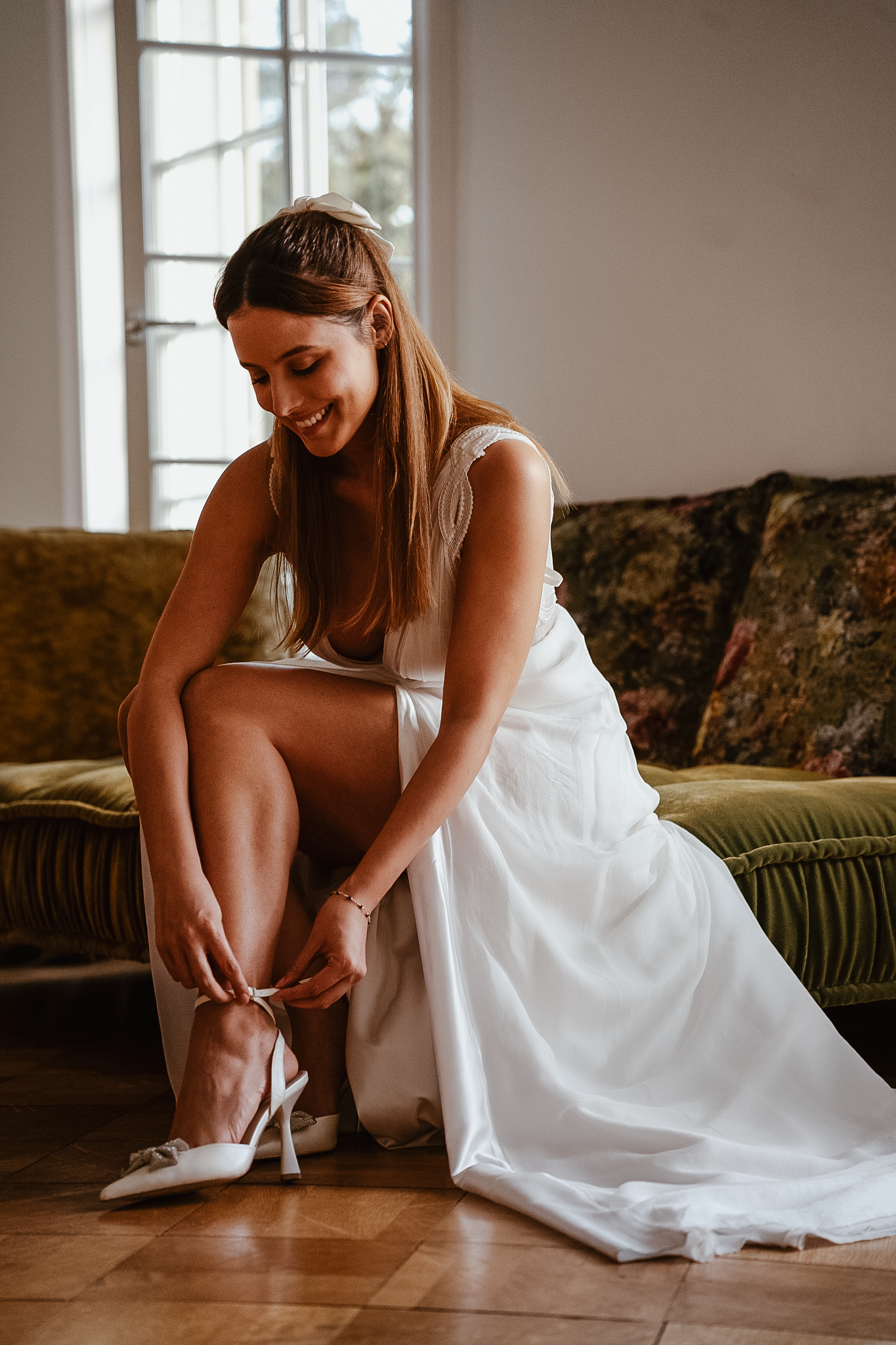 Smiling bride in white wedding dress sitting on green sofa, fastening elegant heeled shoes on wooden floor.