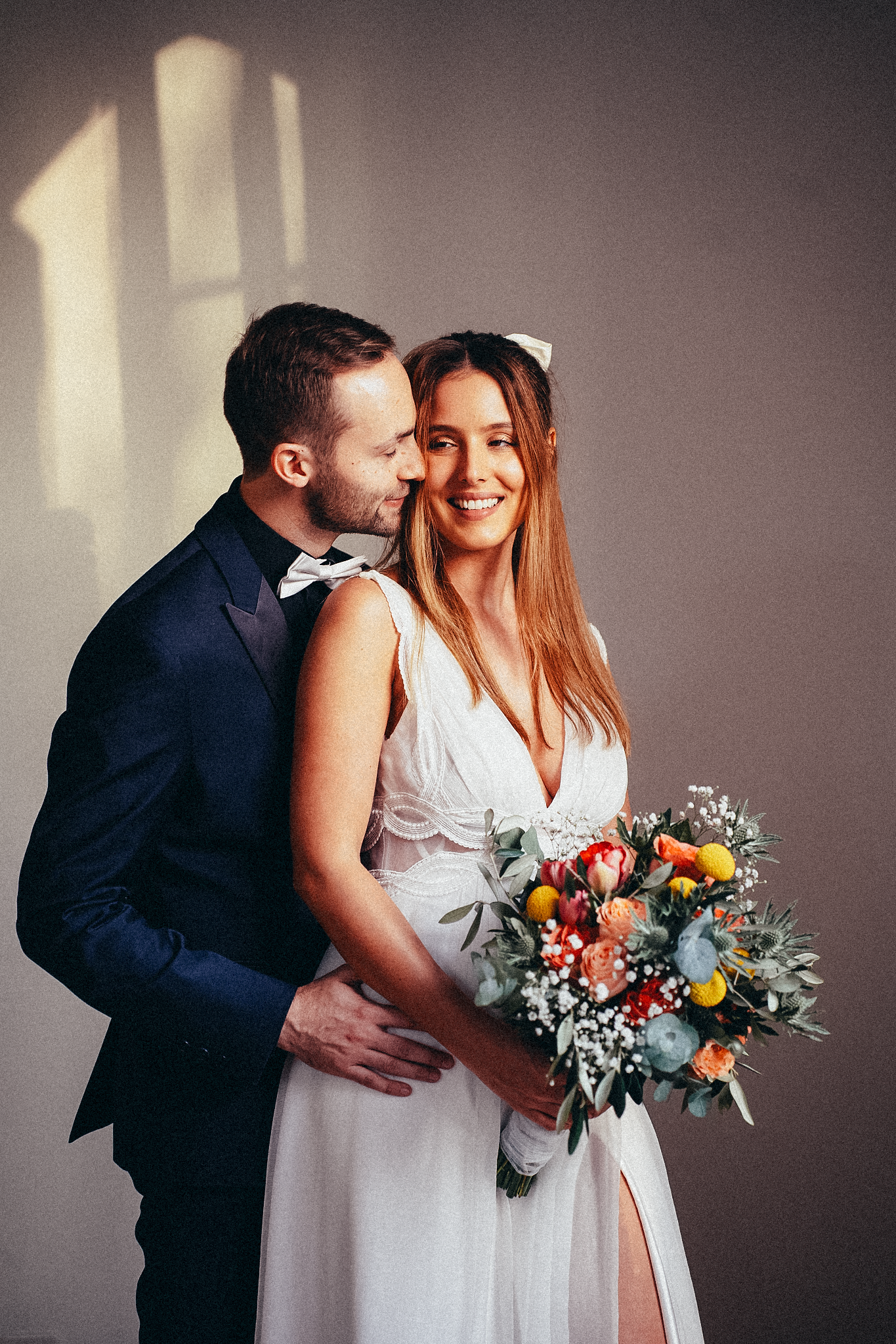 Bride and groom embrace tenderly, bride holding colorful bouquet in white dress, groom in navy tuxedo.