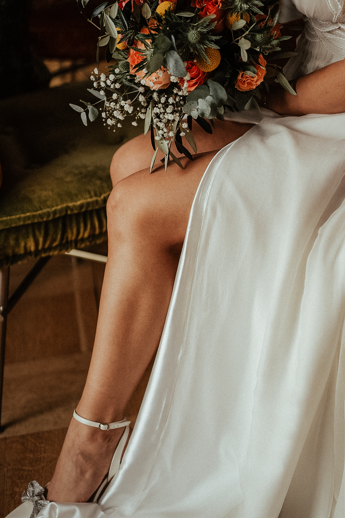 Bride in white gown holding colorful orange and green bouquet, seated with elegant ankle-strap heels.