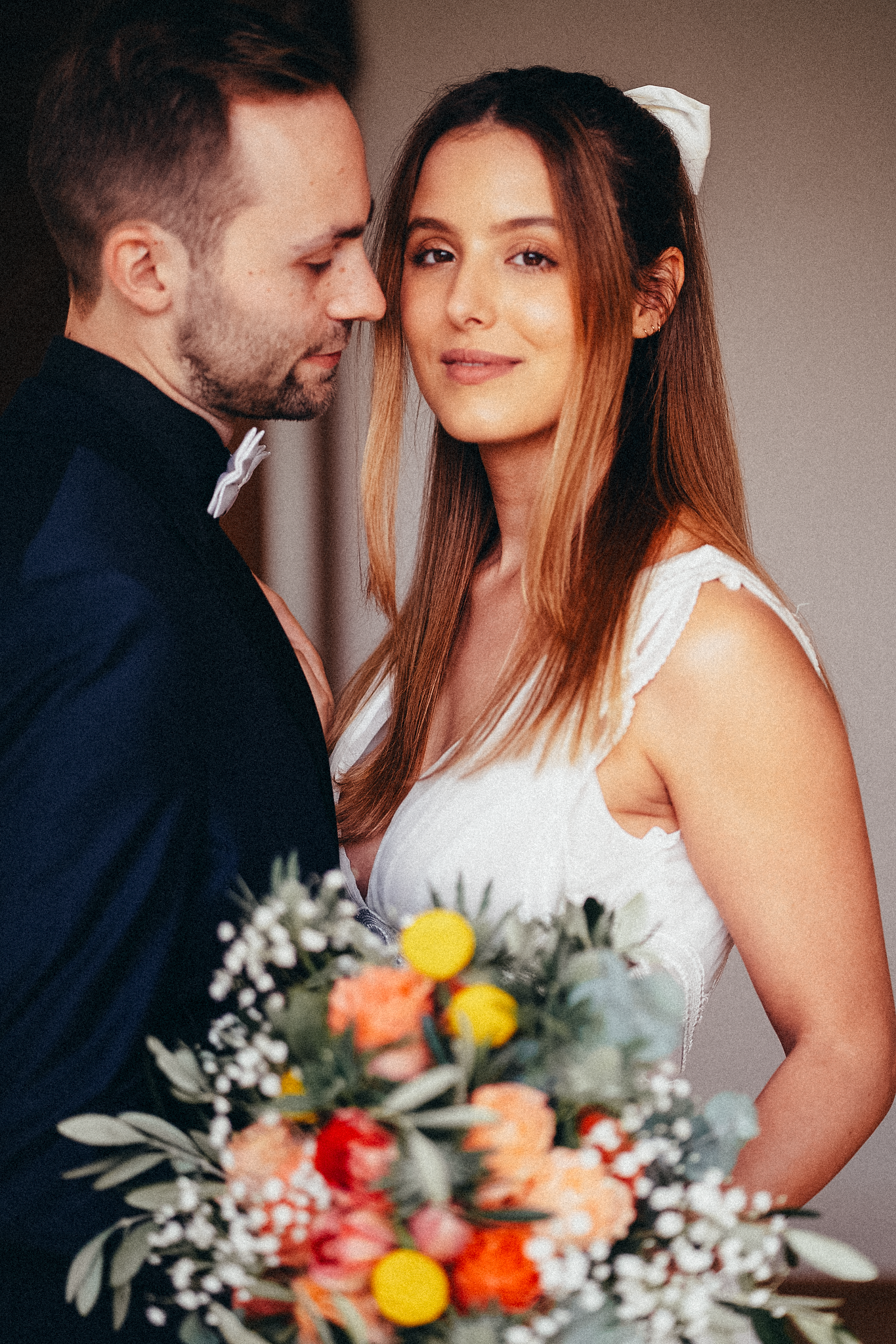 Bride holding colorful bouquet gazes at camera while groom leans in close during intimate wedding portrait.