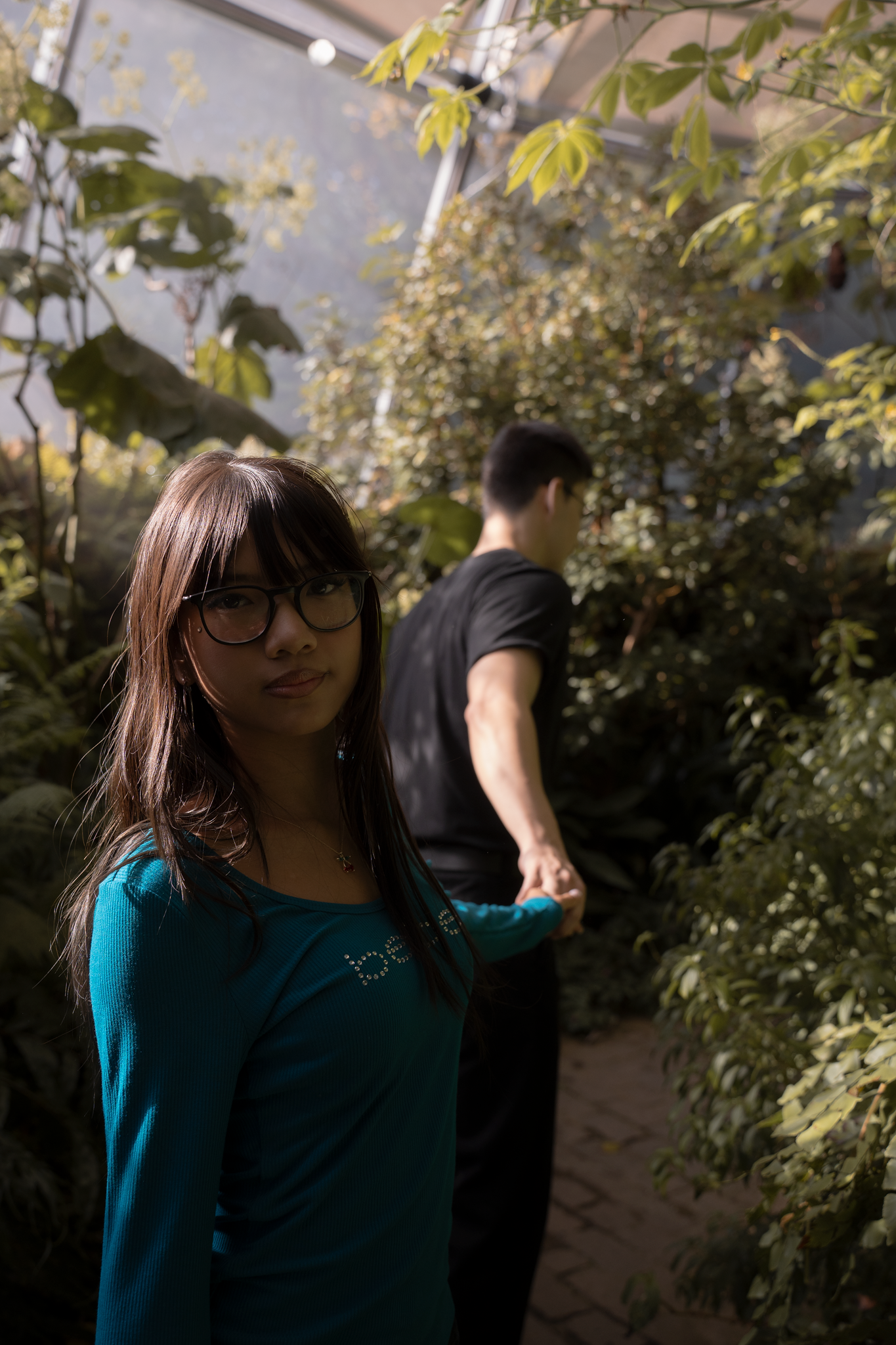 A woman in glasses wearing a teal top stands in a lush, green garden, holding hands with a man in black, evoking a serene and intimate mood.