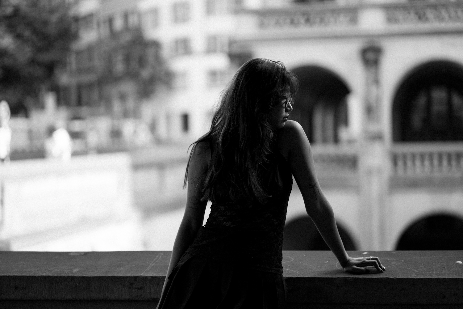 Black and white photo of a woman in a dress leaning on a railing, gazing at a European cityscape.