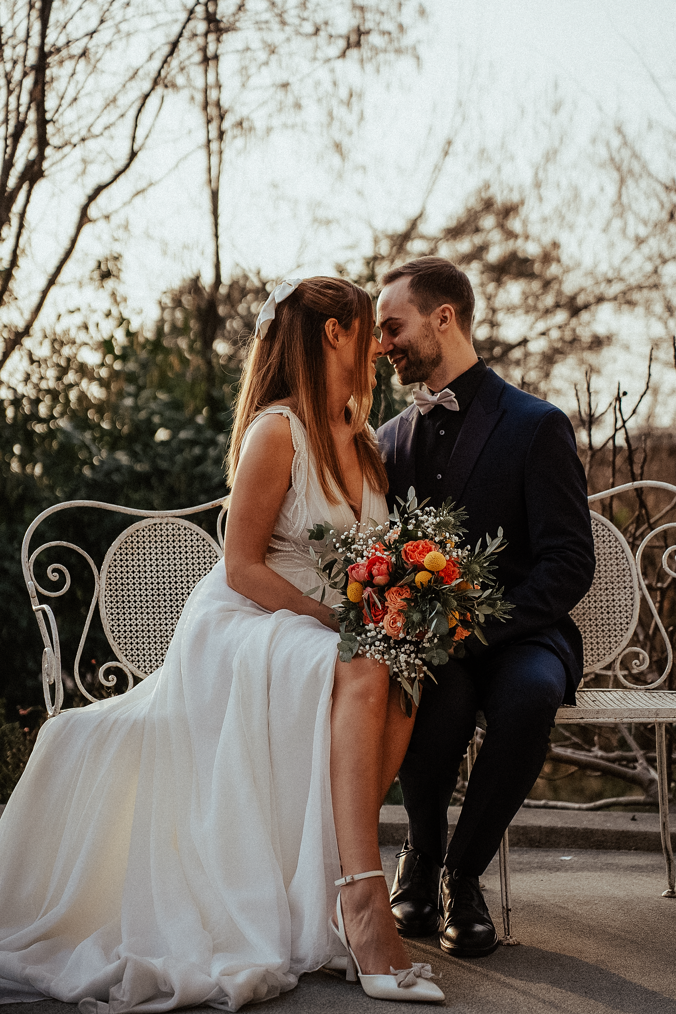 Bride and groom sitting on white iron bench, sharing romantic moment with colorful bouquet, outdoor wedding portrait.