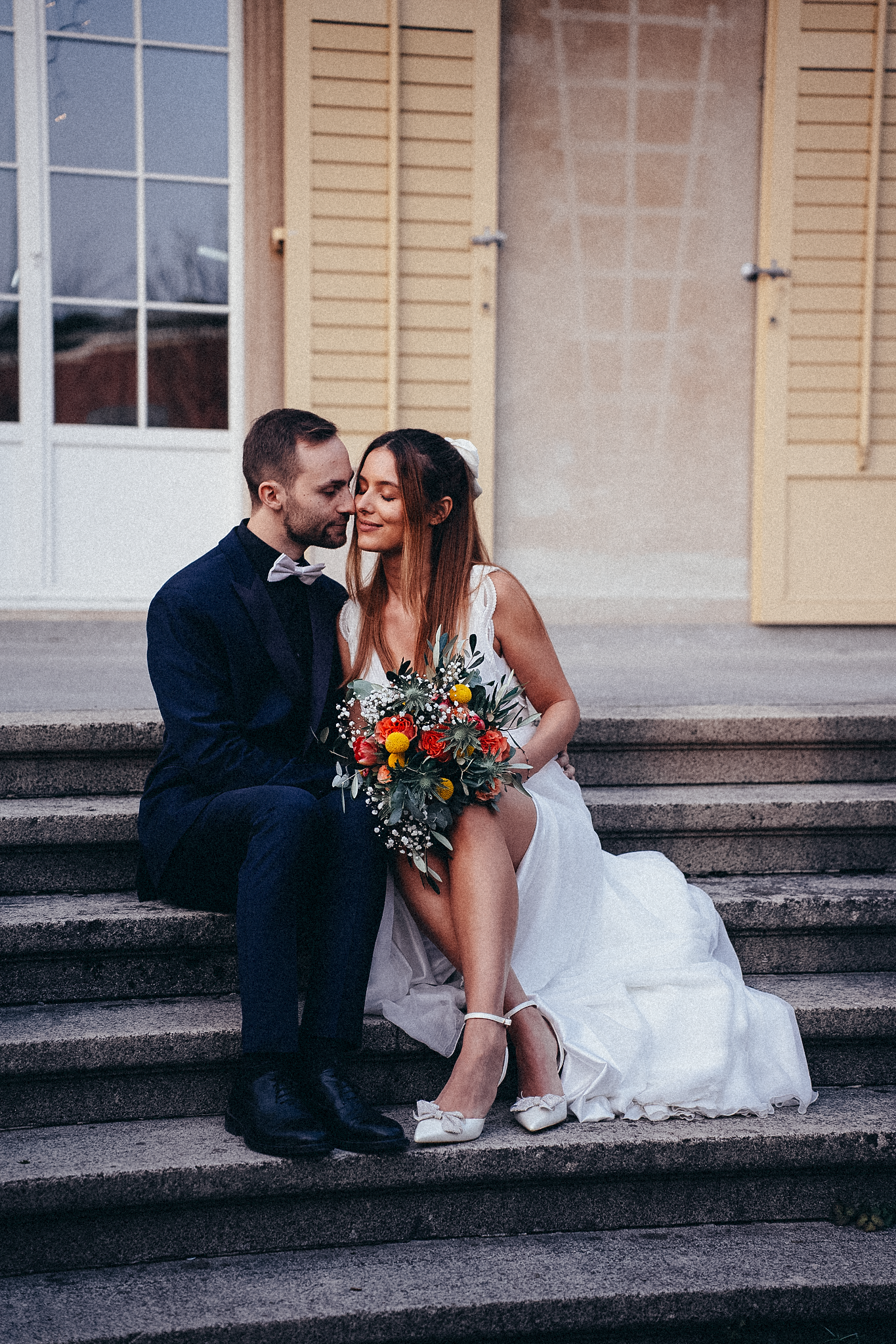 A bride and groom sit intimately on stone steps, she holds a colorful bouquet in a white gown, he wears a navy suit.
