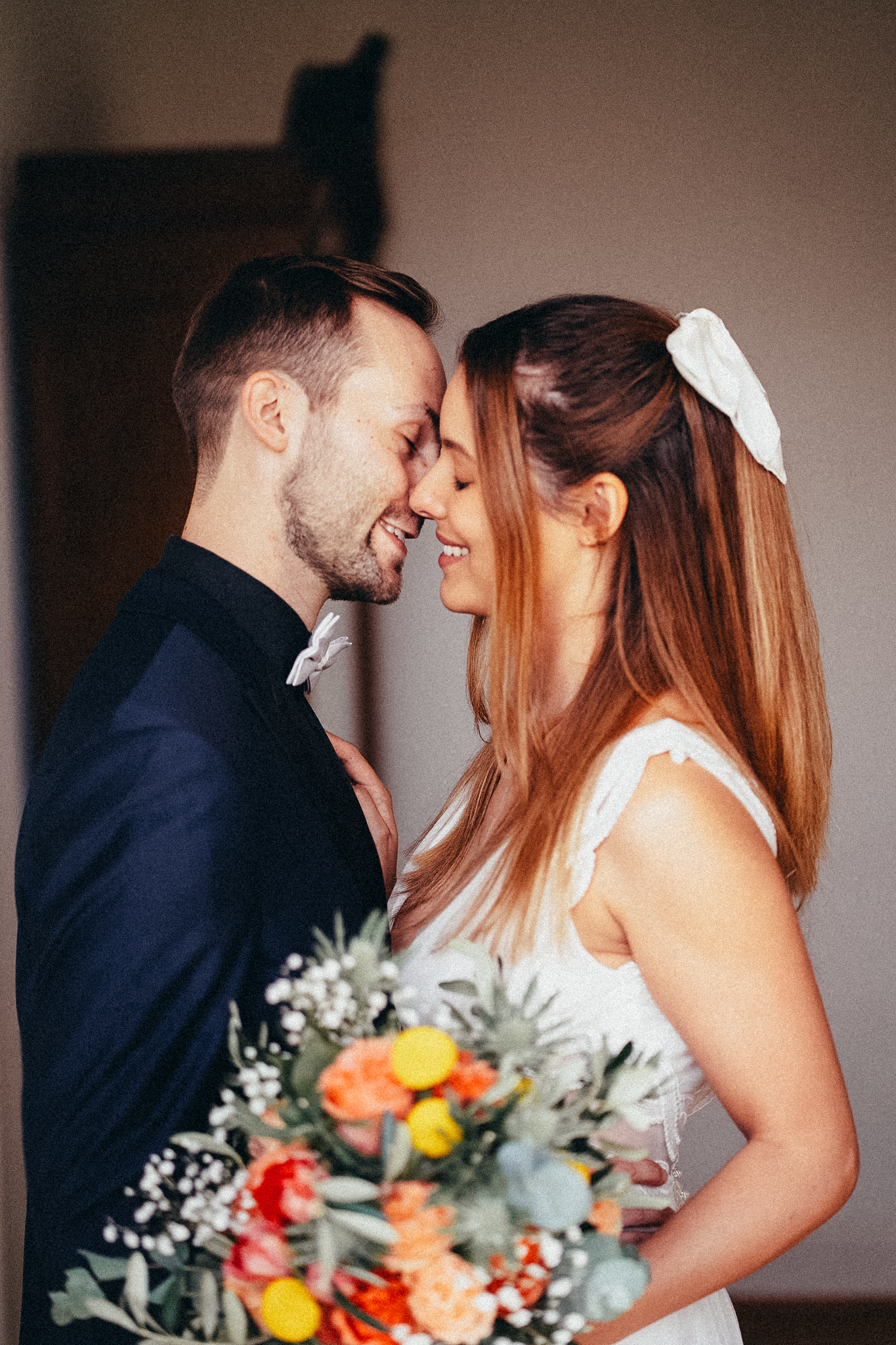 A smiling bride and groom touch foreheads tenderly, she holds a colorful bouquet with orange and yellow flowers on their wedding day.