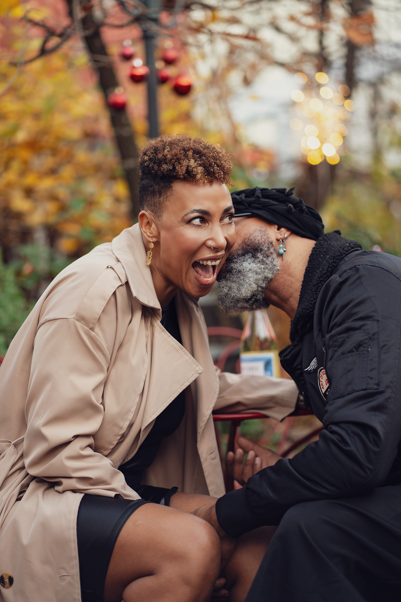 Two people laughing and sharing a joyful moment outdoors in autumn, surrounded by colorful foliage and festive lights.
