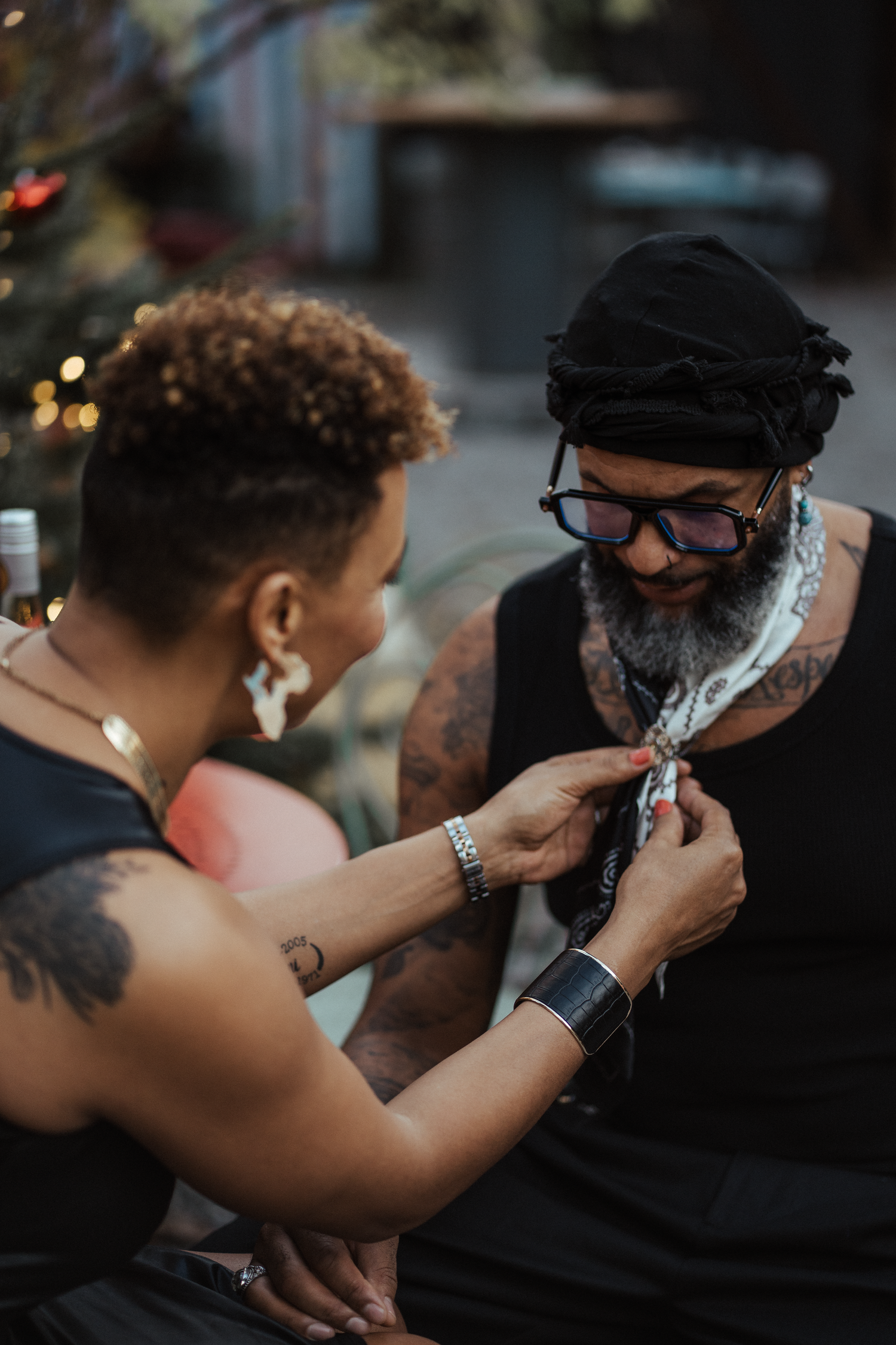 Two tattooed individuals sharing an intimate moment, one adjusting the other's bandana scarf outdoors.