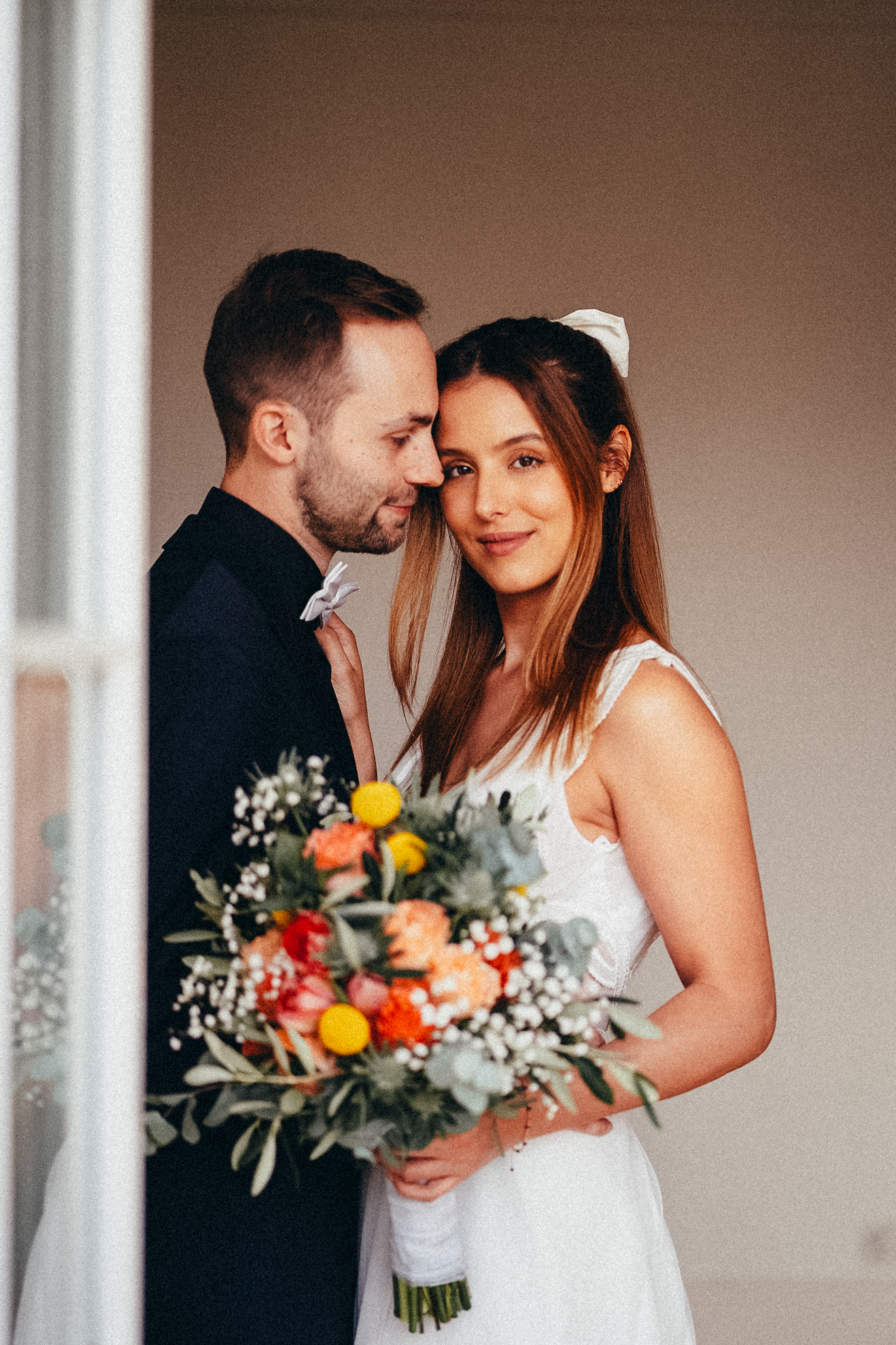 A smiling bride in white dress with bow headband leans close to groom in dark suit holding a colorful bouquet.