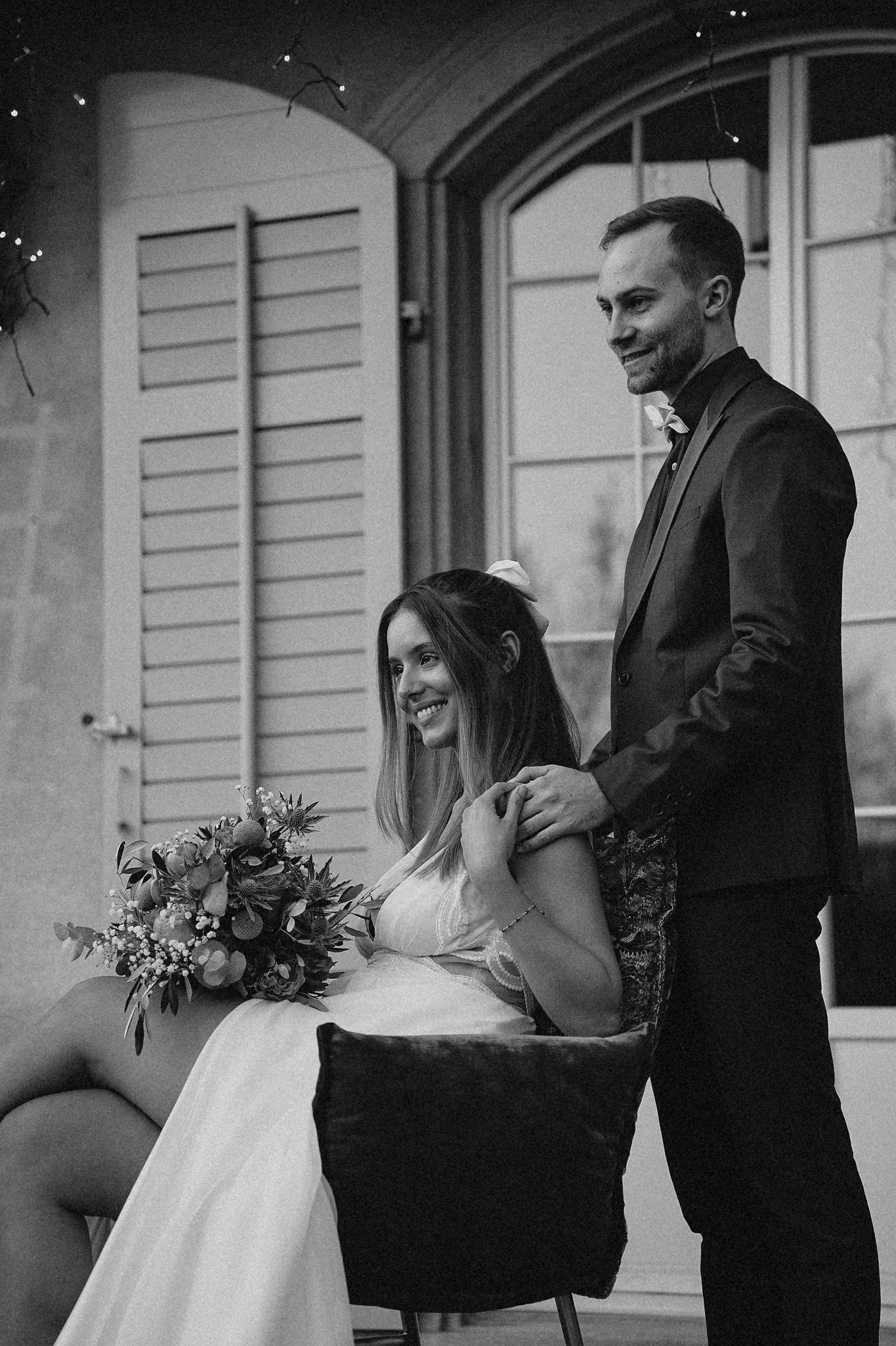 Black and white photo of smiling bride seated holding bouquet while groom stands behind her outdoors.