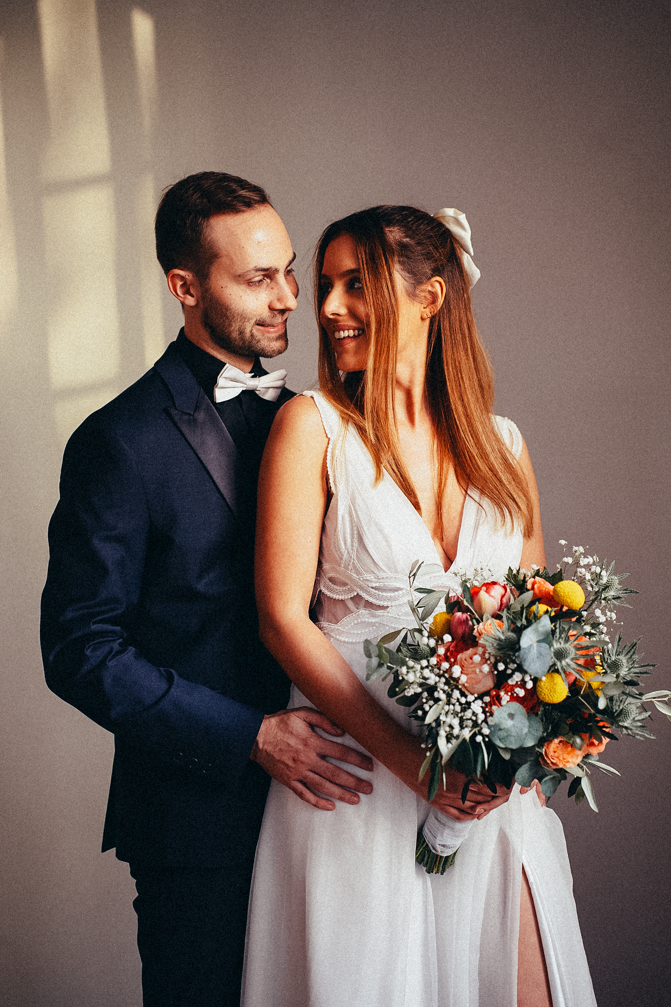 Elegant bride and groom posing indoors, bride holding colorful bouquet in white dress, groom in navy suit with bow tie.