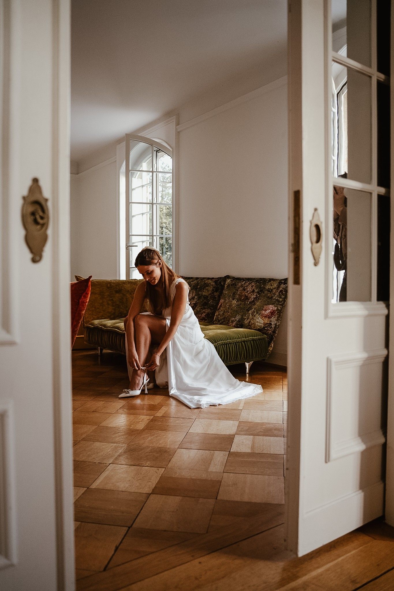 Bride in white wedding dress sitting on floor adjusting her heels in an elegant room with hardwood floors.