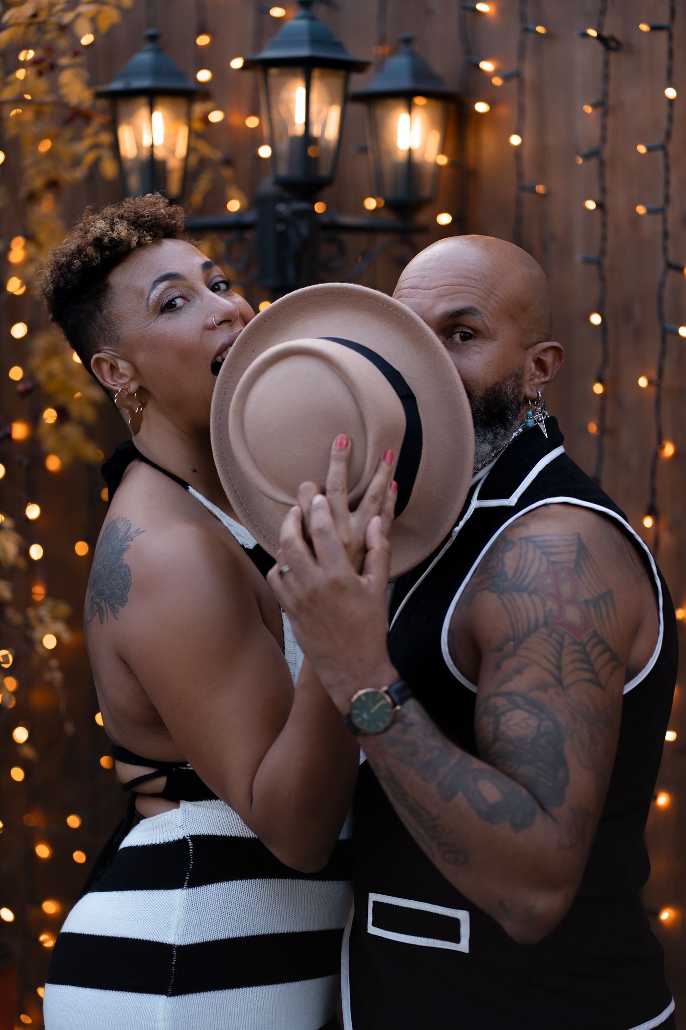 A tattooed couple poses playfully behind a tan hat, surrounded by warm string lights on a wooden backdrop.