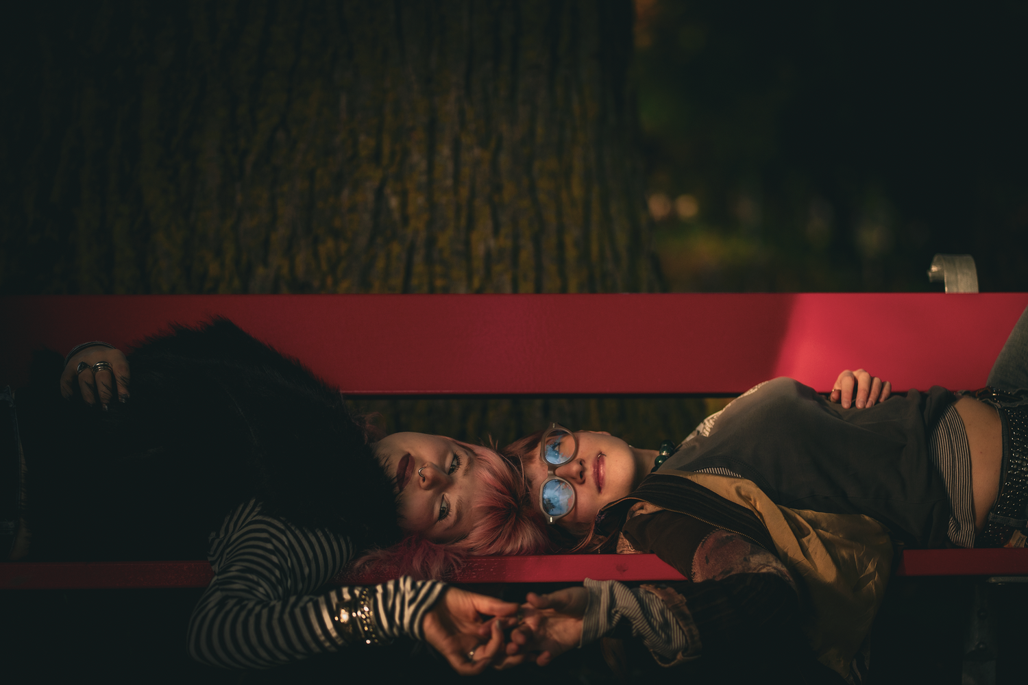 Two people lying on a red bench at night, leaning against each other near a large tree.