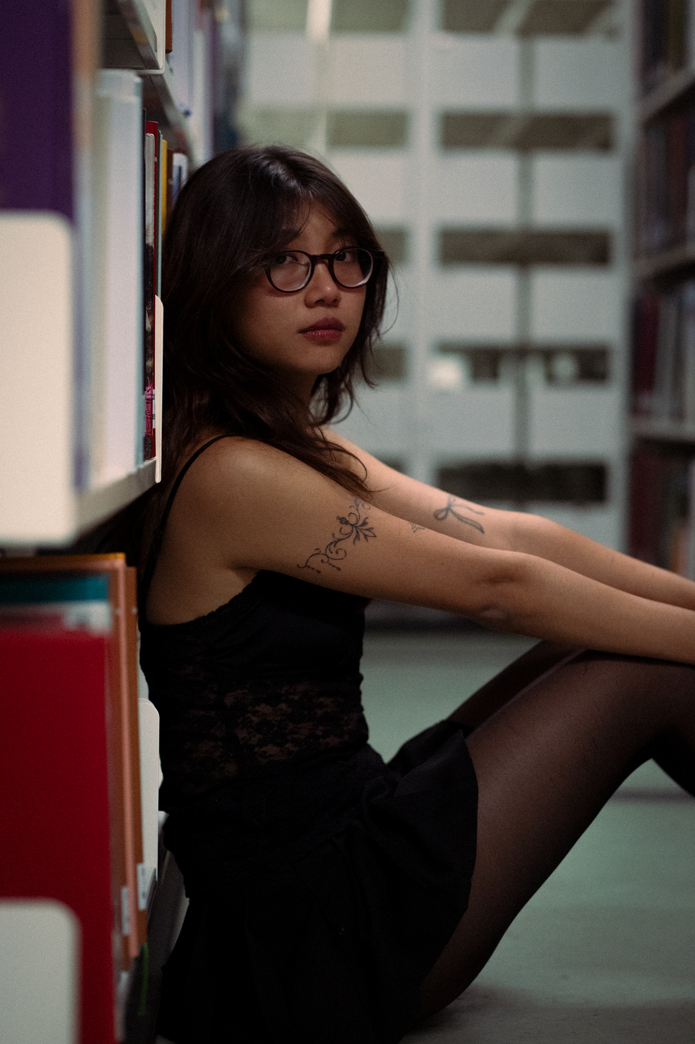 Young woman with glasses and arm tattoo in black dress sits against library bookshelf on floor.