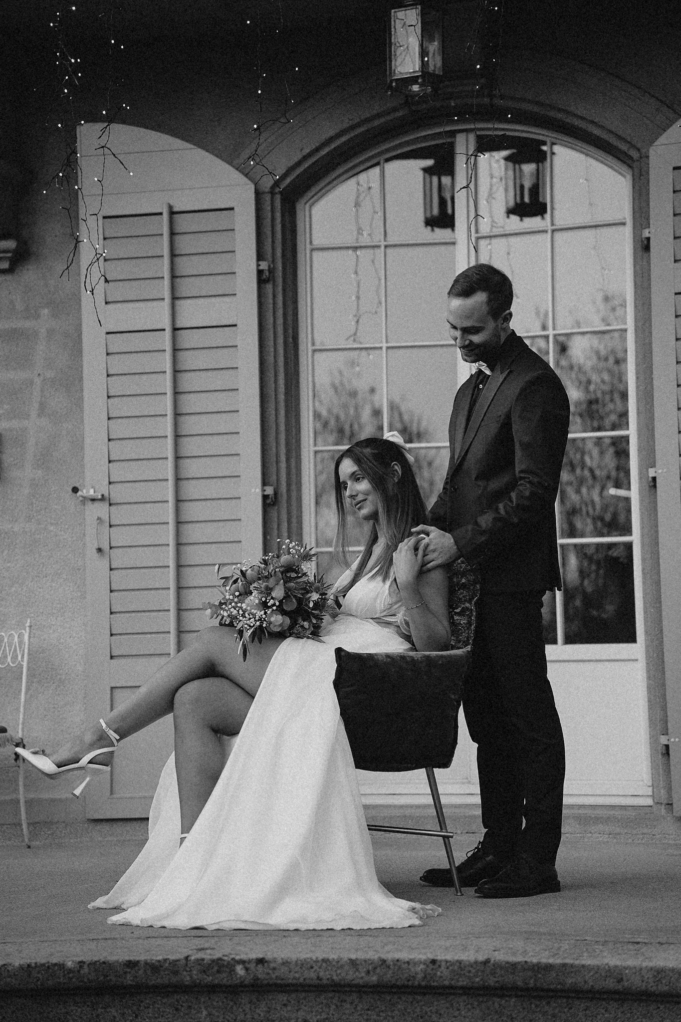 Elegant black and white wedding portrait of seated bride holding bouquet with groom standing beside her.