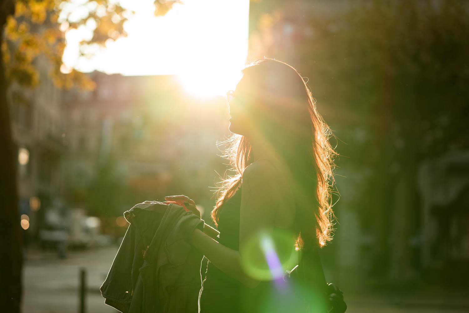Silhouetted woman with flowing hair stands on a sunlit street, holding a jacket. The bright sunlight creates a warm and serene atmosphere.