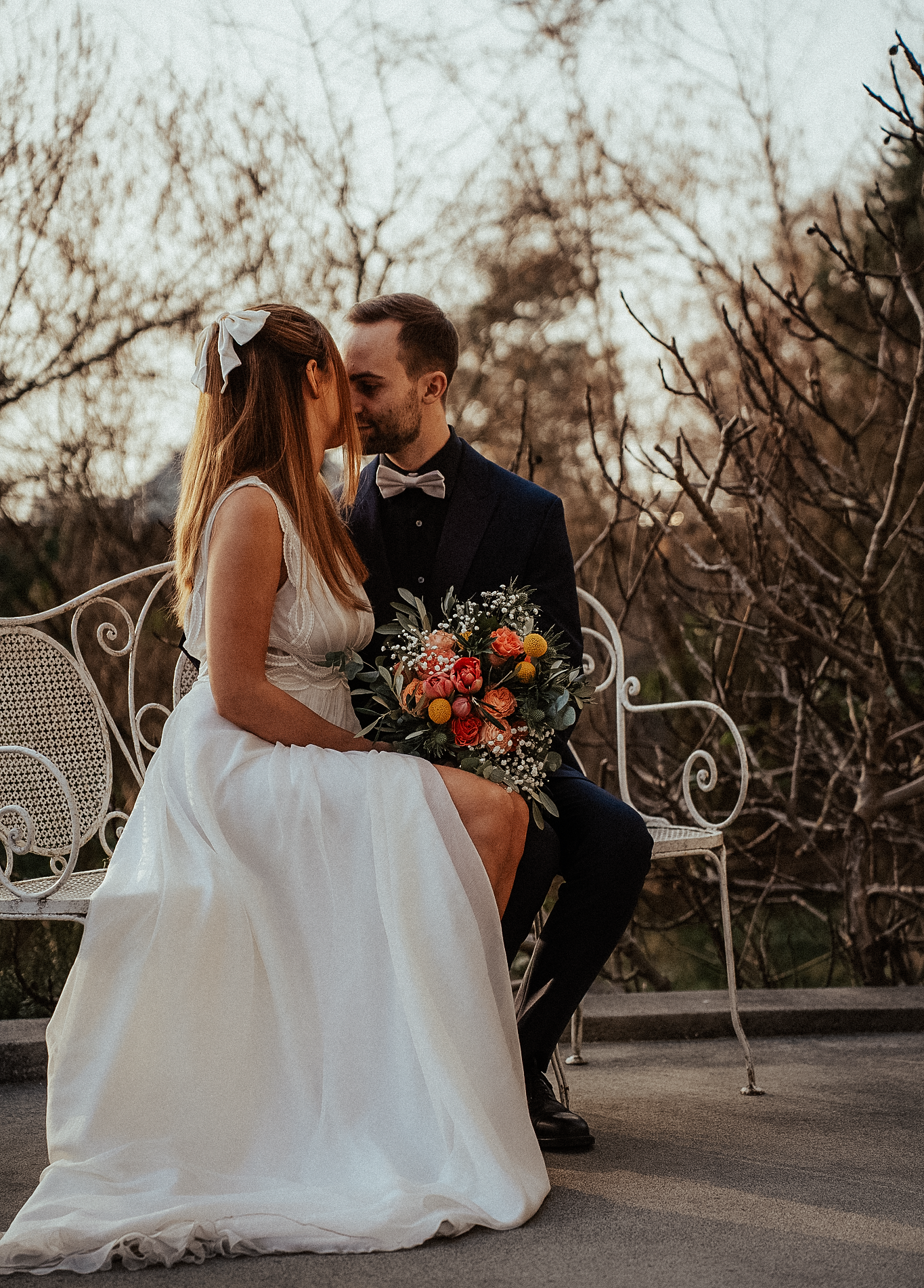 Bride and groom share a tender kiss on decorative white iron bench surrounded by winter bare trees.