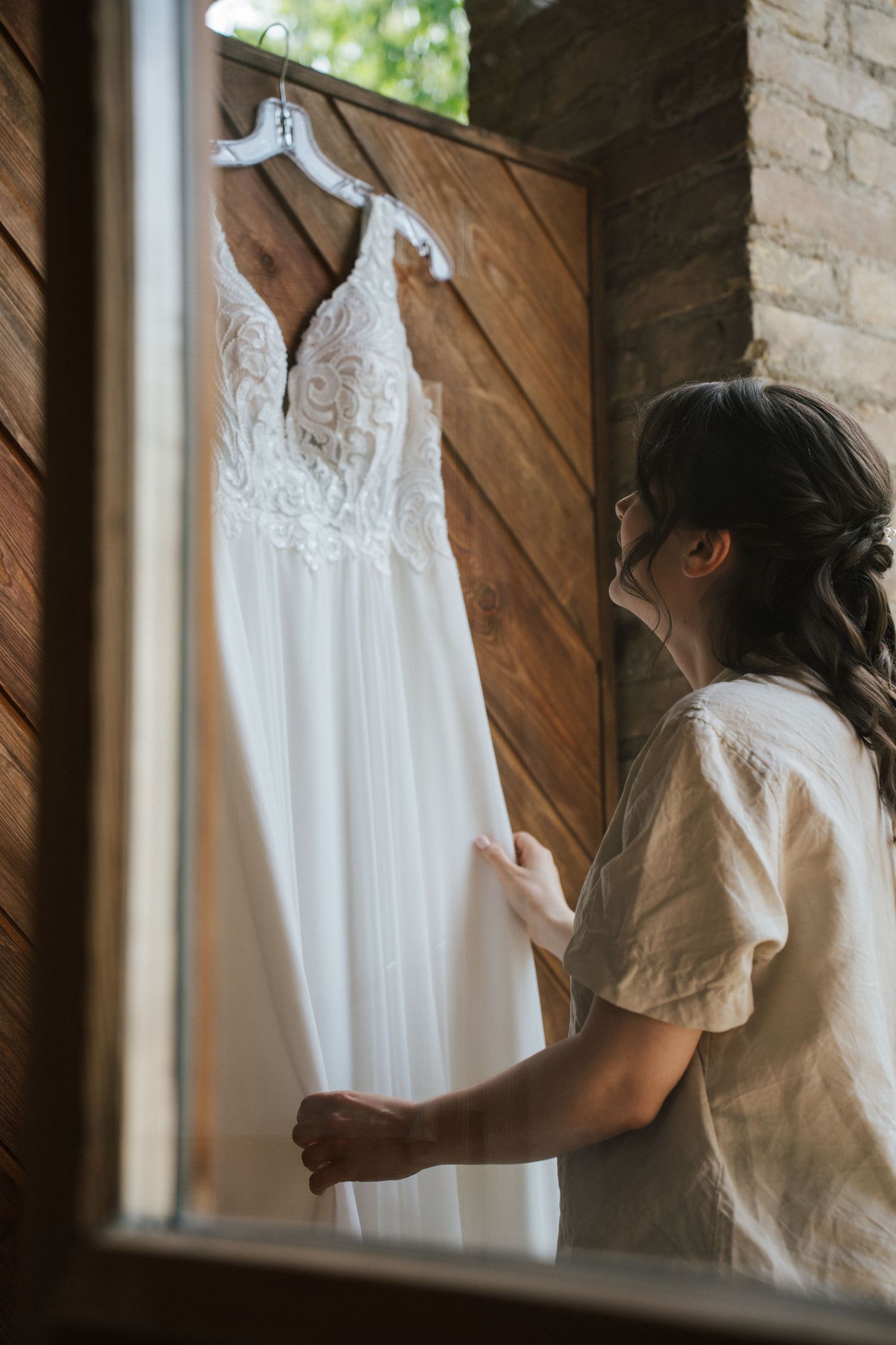 Bride admiring her white lace wedding dress hanging on a wooden door in a rustic setting.
