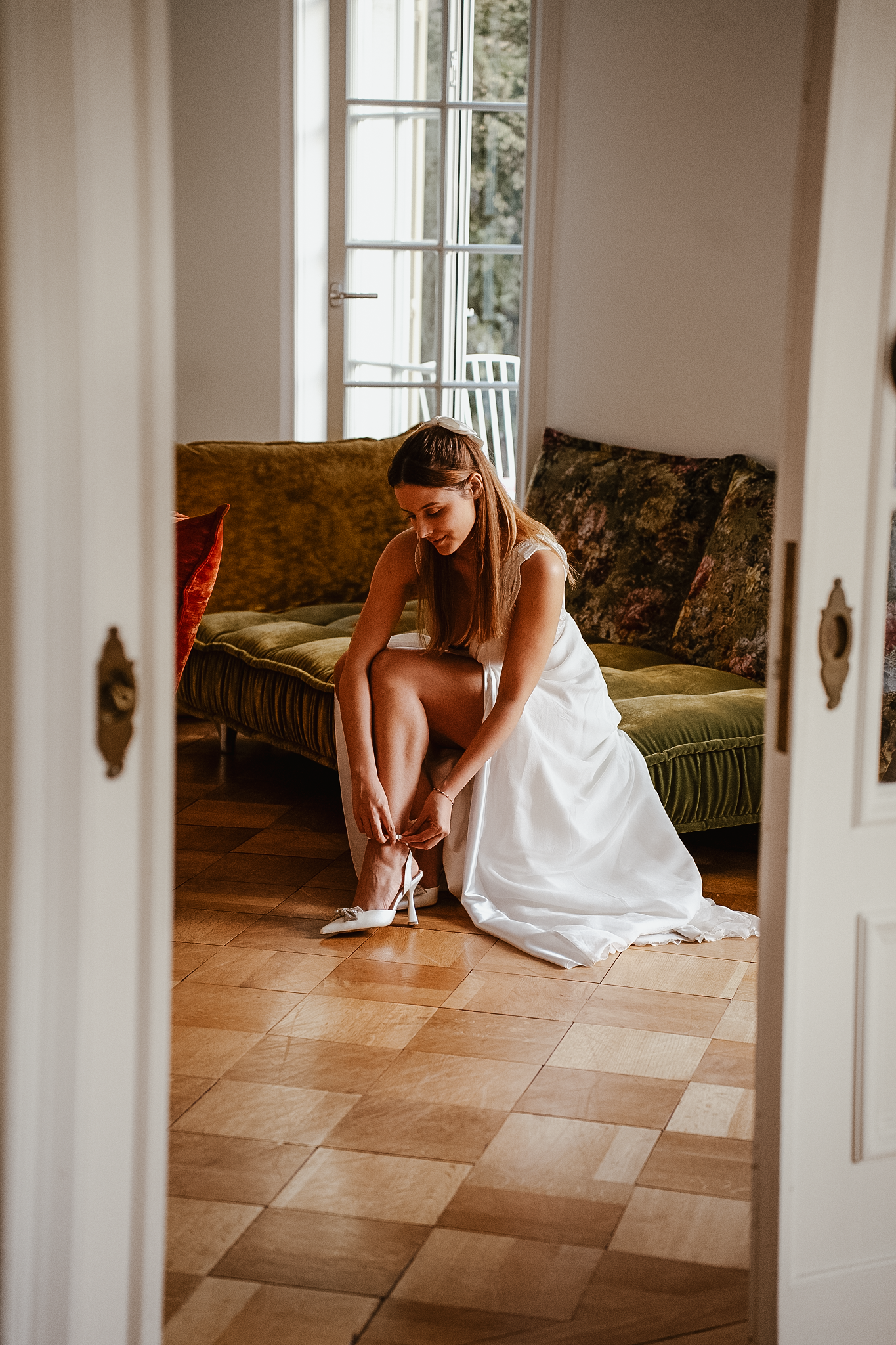 Bride in elegant white wedding dress sitting on green sofa putting on heels in a sunlit room.