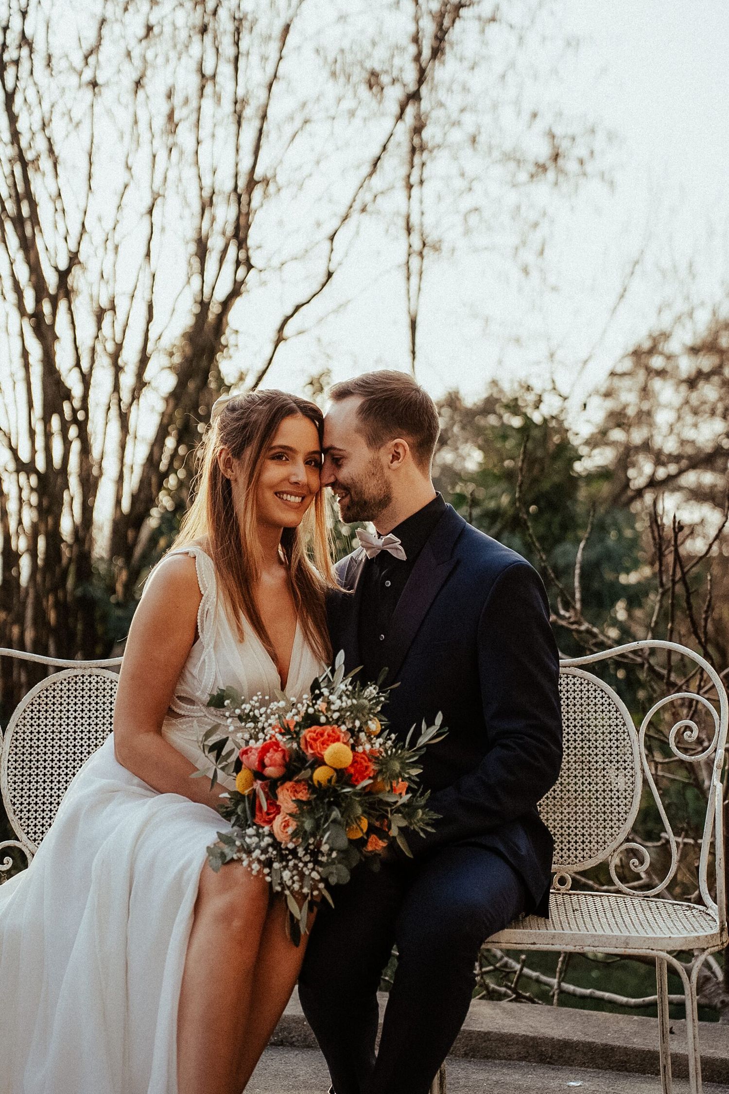 Bride and groom sitting on white iron bench, sharing romantic moment with colorful bouquet, trees in background.