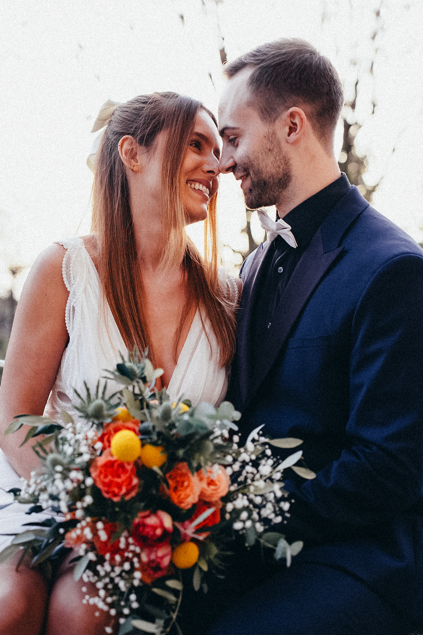 Smiling bride and groom touch foreheads tenderly, bride holding a colorful wedding bouquet outdoors.