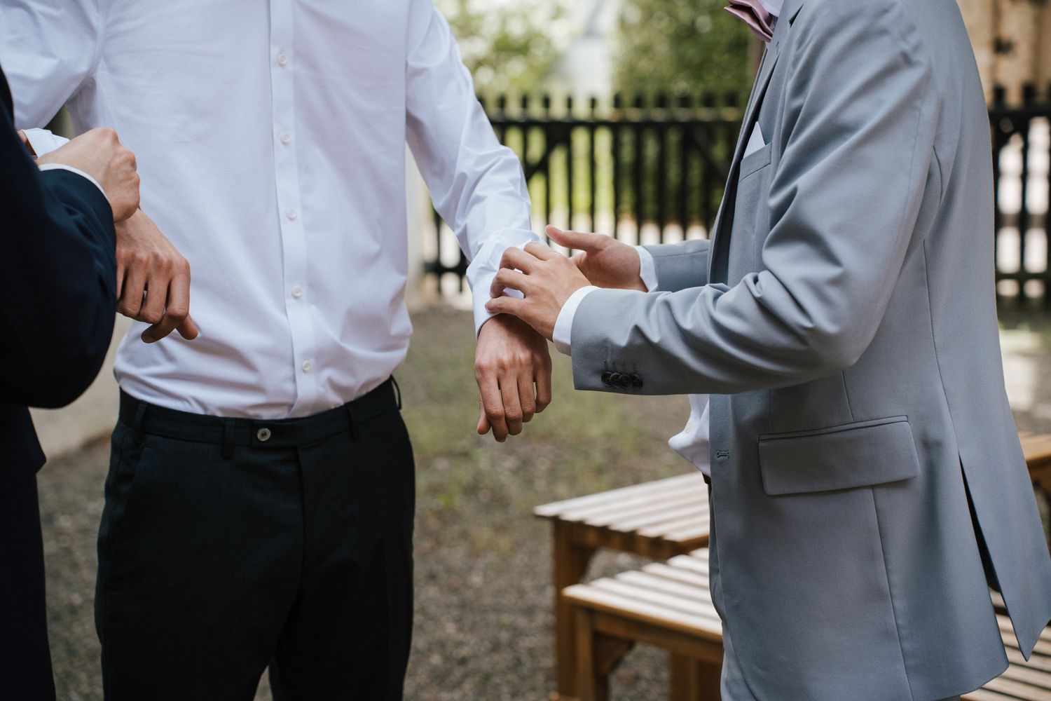 Man in gray suit adjusting cufflinks on groom in white shirt outdoors at wedding ceremony.