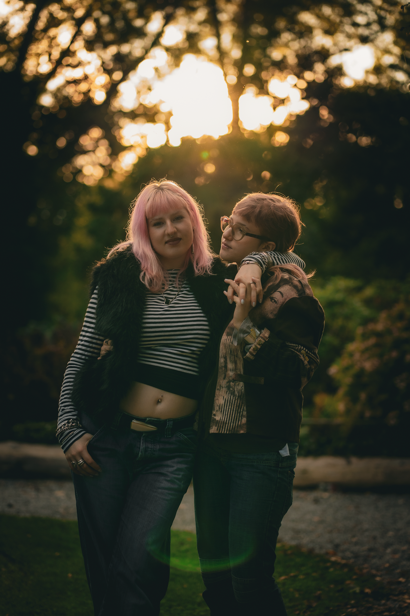 Two people stand closely together outdoors at sunset, one with pink hair and striped shirt, the other with glasses and casual attire.
